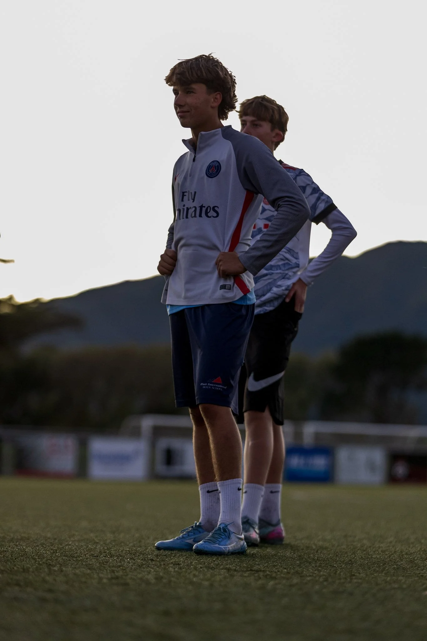 Young man standing on a sports field, facing away, wearing a black t-shirt that says 'IT STARTS HERE' and black shorts, with a chain necklace, with a chain link fence and mountain landscape in the background.