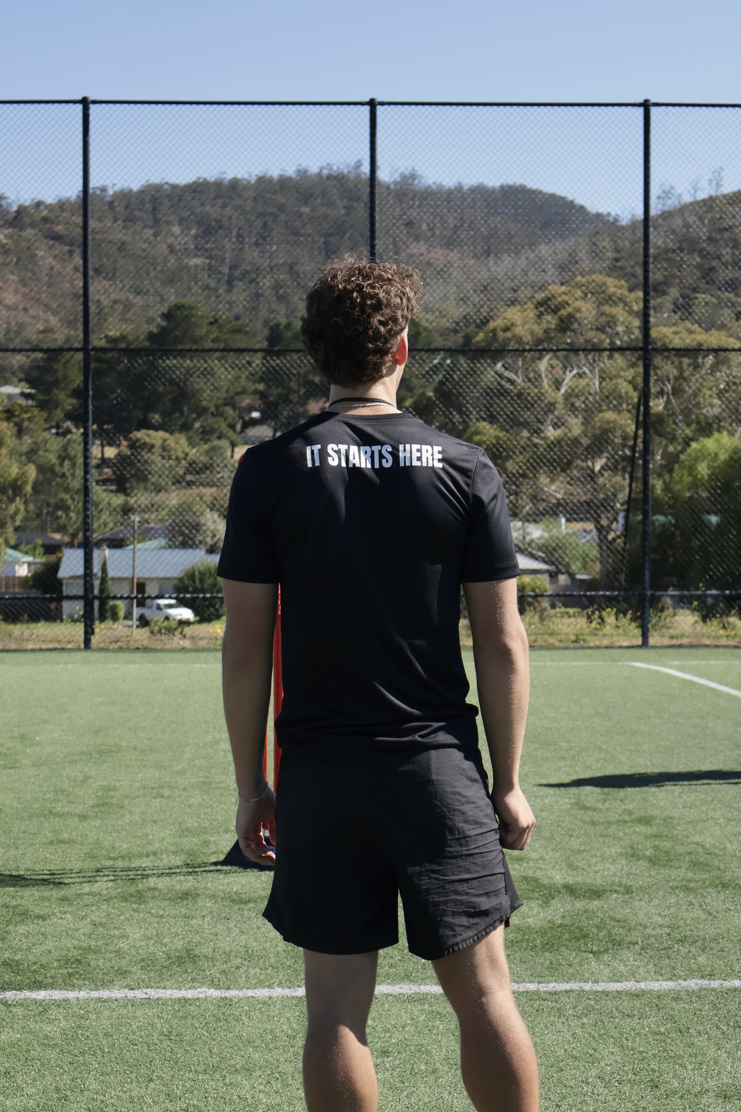 Young man standing on a sports field, facing away, wearing a black t-shirt that says 'IT STARTS HERE' and black shorts, with a chain necklace, with a chain link fence and mountain landscape in the background.