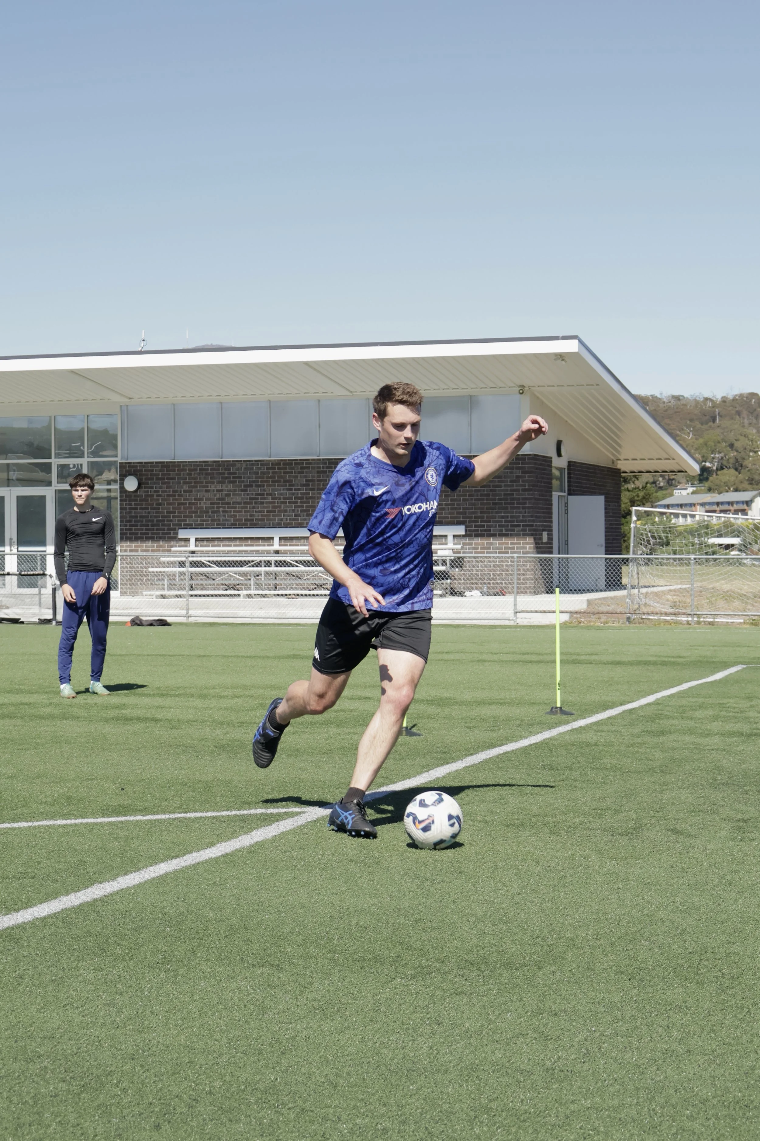 Young man in blue soccer jersey running on a soccer field with a soccer ball, with another person observing in the background.