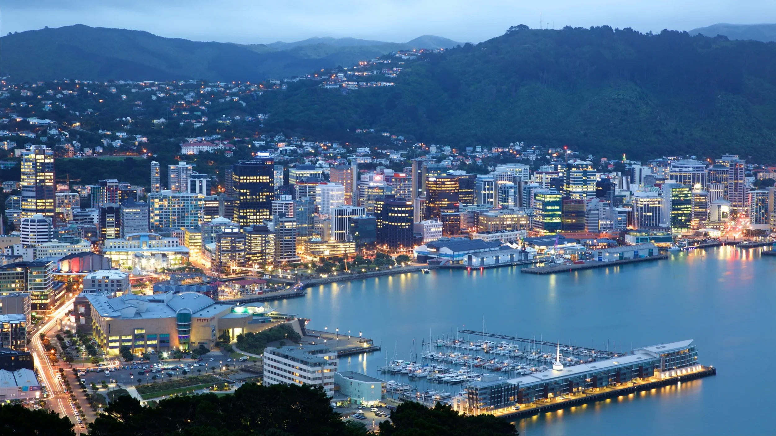 A city skyline at dusk with illuminated buildings, a harbor with boats, and mountains in the background.