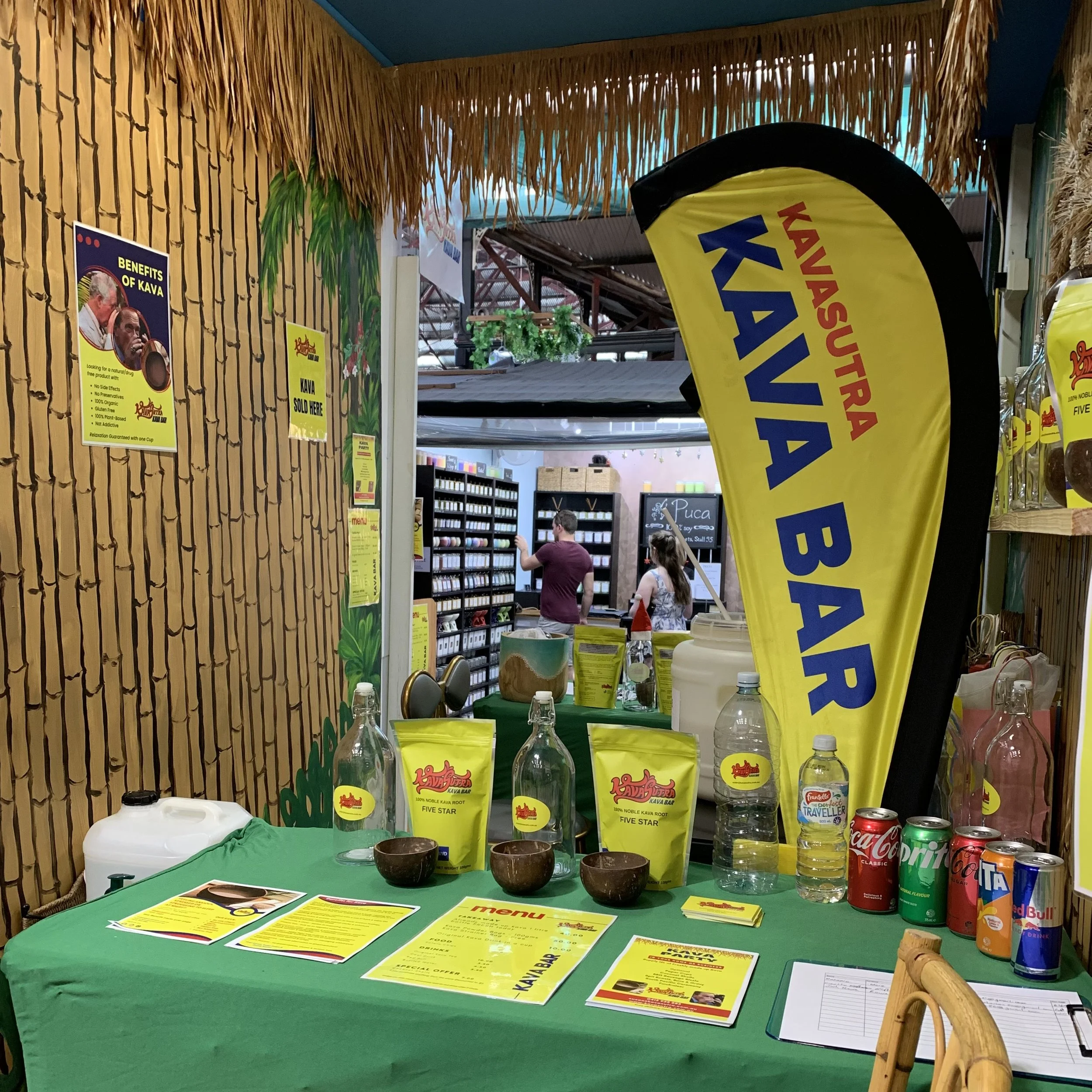 Display table at a Kava Bar with promotional materials, bags of Kava, bottles of water, soda cans, and a large yellow Kava Bar flag. In the background, a couple of people are browsing shelves.