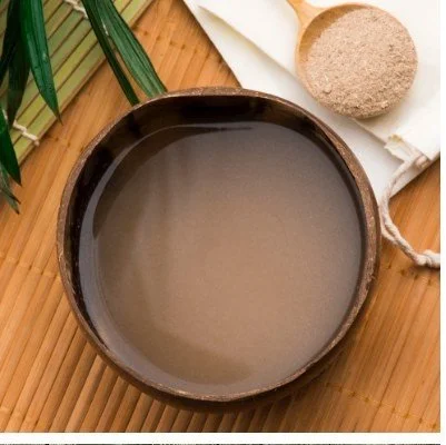 A bowl of liquid on a wooden surface with a loofah and some green leaves nearby.