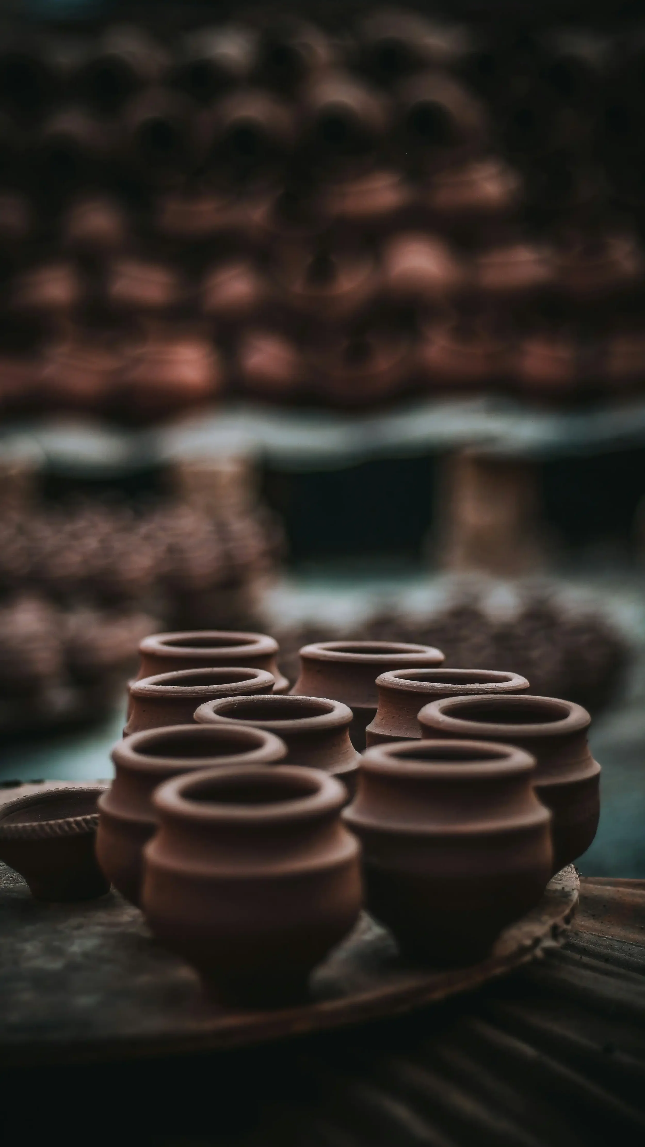 Unbaked clay pots arranged on a tray, with a background of blurred additional pottery in a pottery workshop.