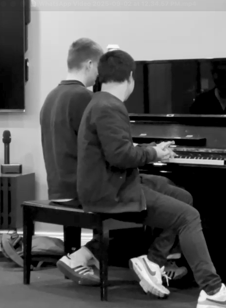 Joel and Luke playing piano together in a room, sitting on a bench in front of the piano.