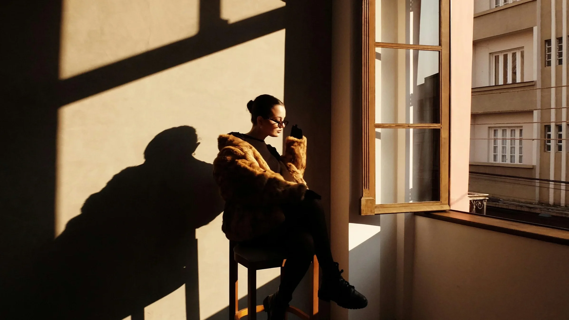 A woman wearing glasses and a fur coat sitting on a stool near an open window, with warm sunlight casting shadows on the wall behind her.