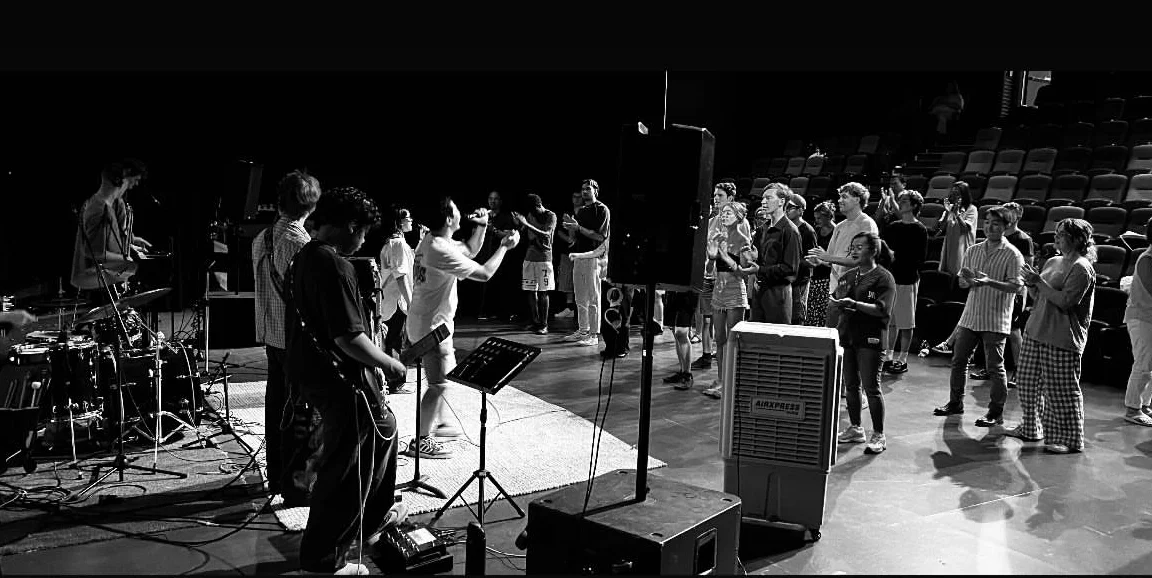 A group of people standing in a music session, some with musical instruments, others holding sheets or music stands, engaged in singing or playing music, with a band or group of musicians leading them in worship. Held at Atelier Theatre, Mount Barker
