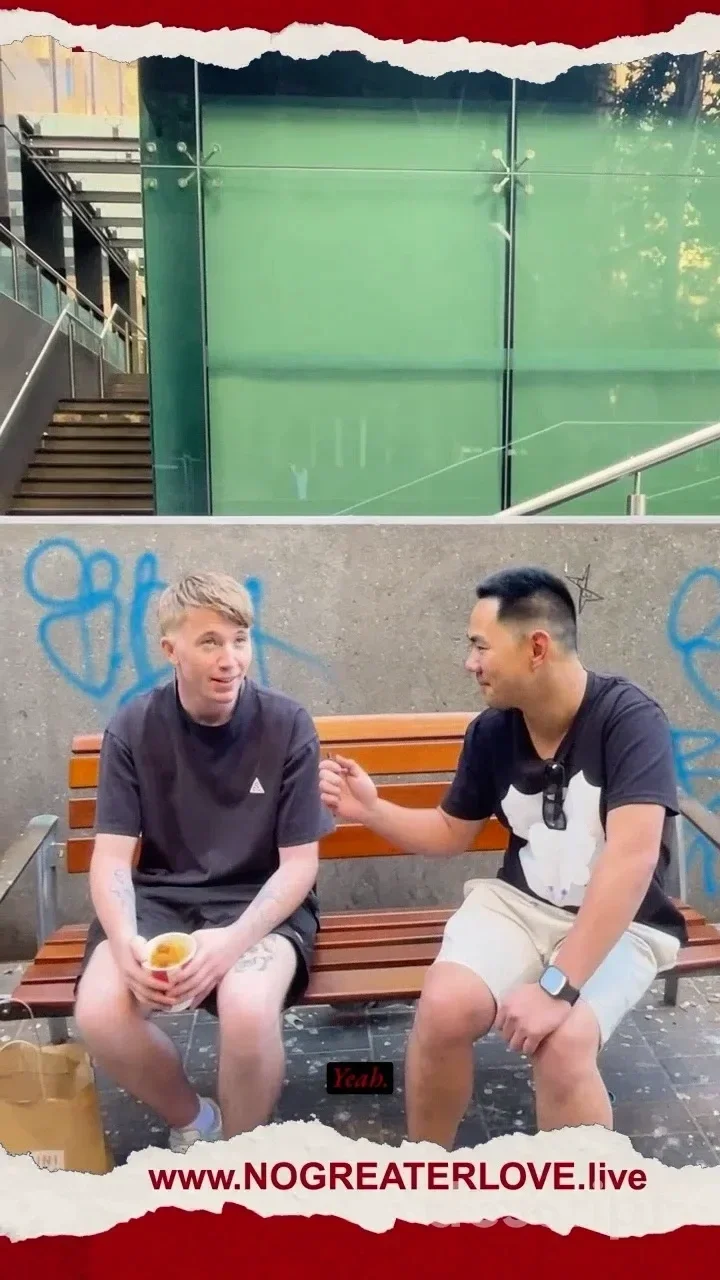 Two young men sit on a bench in a public space. One of them holds a KFC GoBucket while the other holds a portable microphone. There is a URL art the bottom for www.NOGREATERLOVE.live.