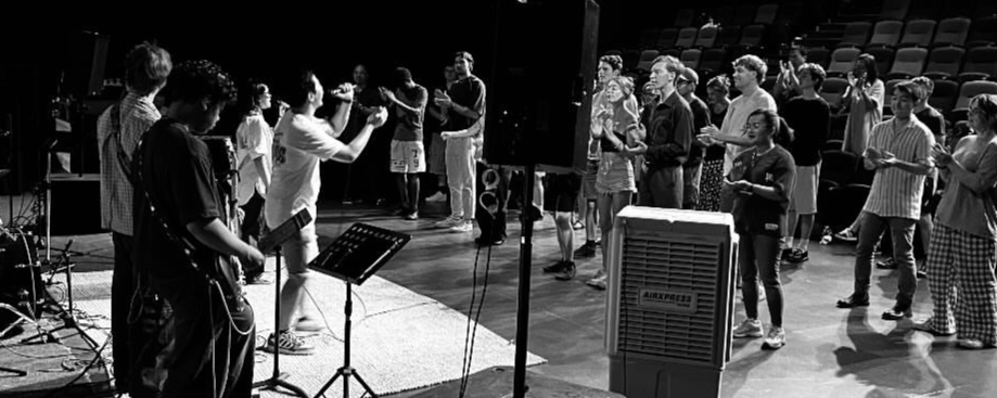 A group of people standing in a music session, some with musical instruments, others holding sheets or music stands, engaged in singing or playing music, with a band or group of musicians leading them in worship. Held at Atelier Theatre, Mount Barker