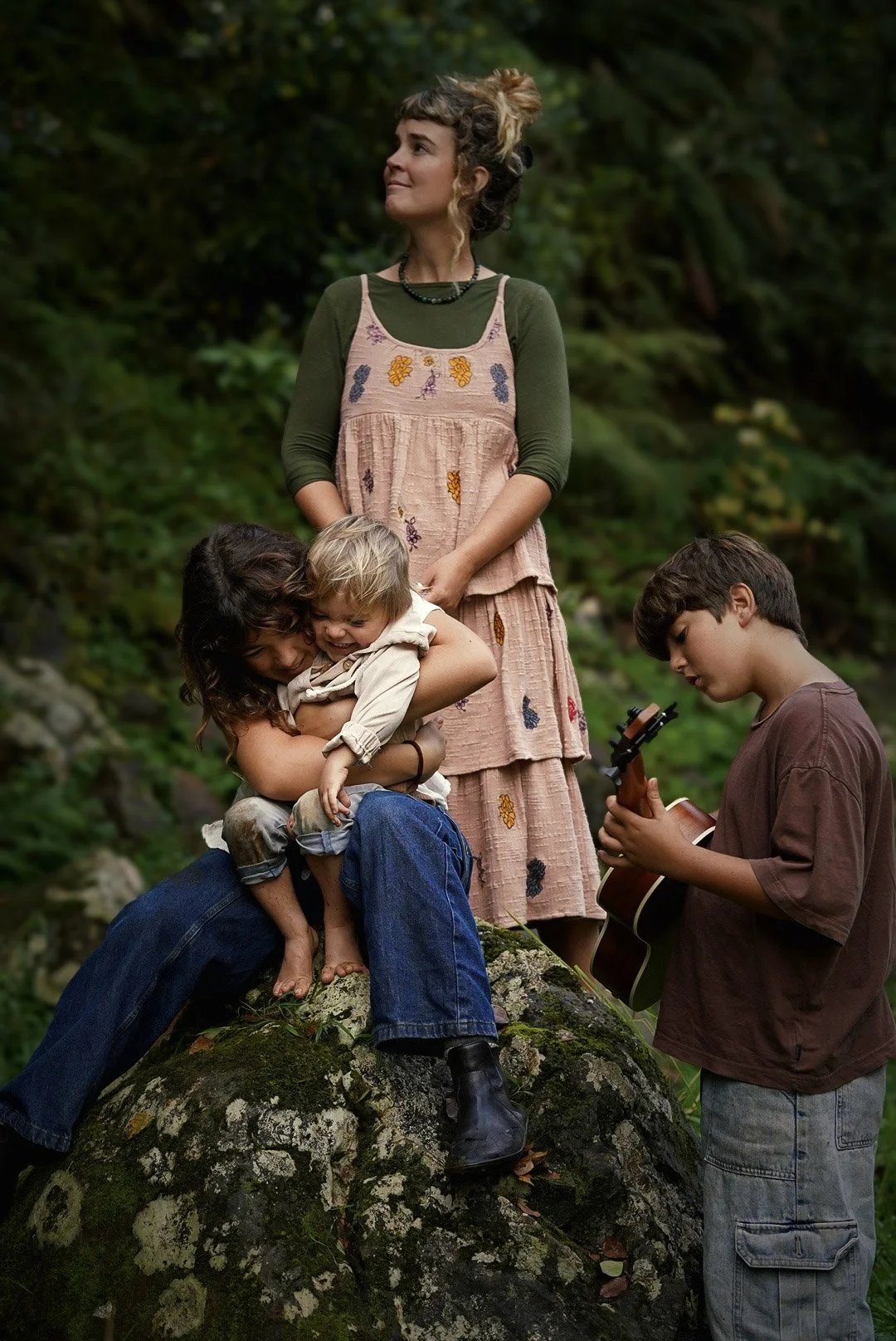 A woman and three children are enjoying time outdoors. The woman is standing with two children, hugging and sitting on a mossy rock, while a boy is playing guitar nearby. Family.