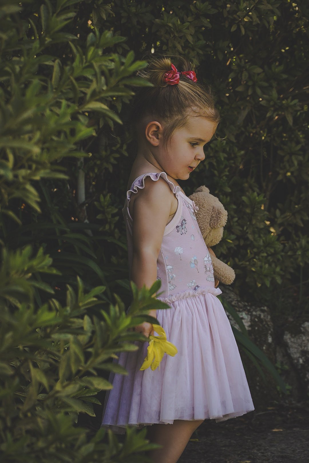 A young girl in a pink dress holding a teddy bear and yellow flower, standing among greenery, looking down thoughtfully.