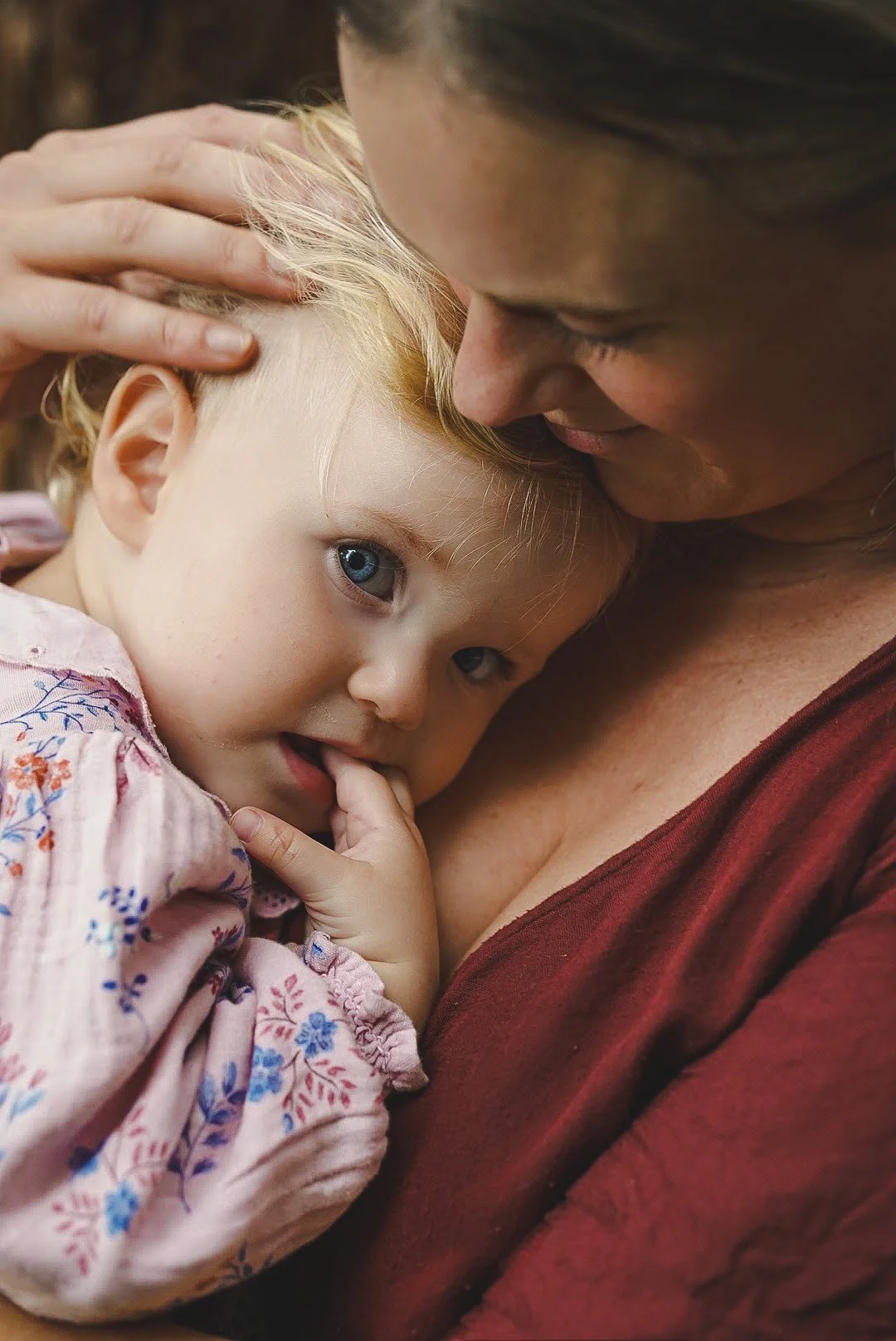A young girl with blue eyes and blonde hair is being held closely by her mother, who is gently touching her head. The girl is resting her head on the woman's chest and has a finger in her mouth, with a calm and affectionate expression.