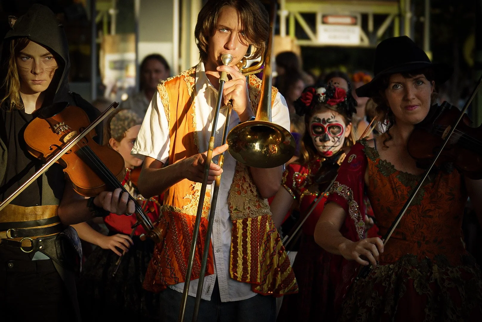 Group of people dressed in costumes, playing musical instruments, with one person in traditional Mexican Day of the Dead face paint at a festive outdoor event.