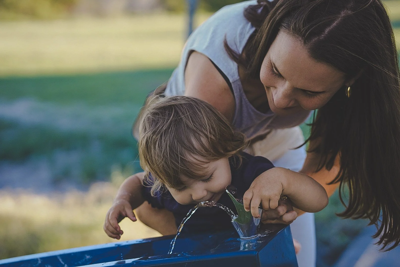 A young child is drinking water from a blue outdoor water fountain while being held by his mother in a park setting.