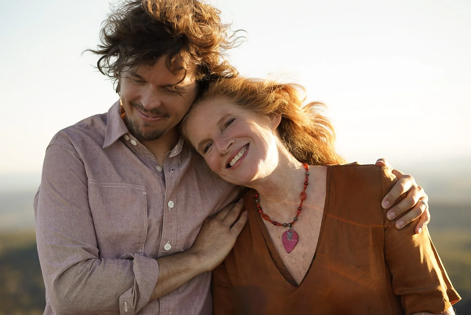 A middle-aged woman and a young man smiling and hugging outdoors during sunset.