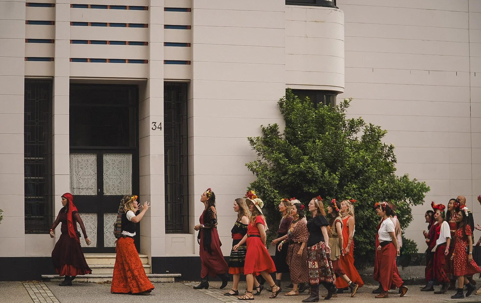 A group of people dressed in colourful, traditional Spanish clothing walking in a line outside a modern building with a large tree nearby.