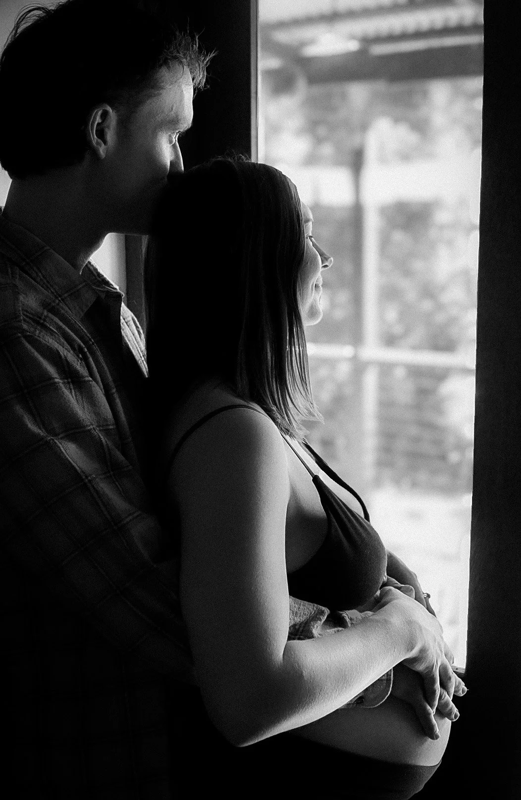 A black-and-white photo of a man and woman standing close by a window, with the man gently embracing the pregnant woman, both looking out the window.