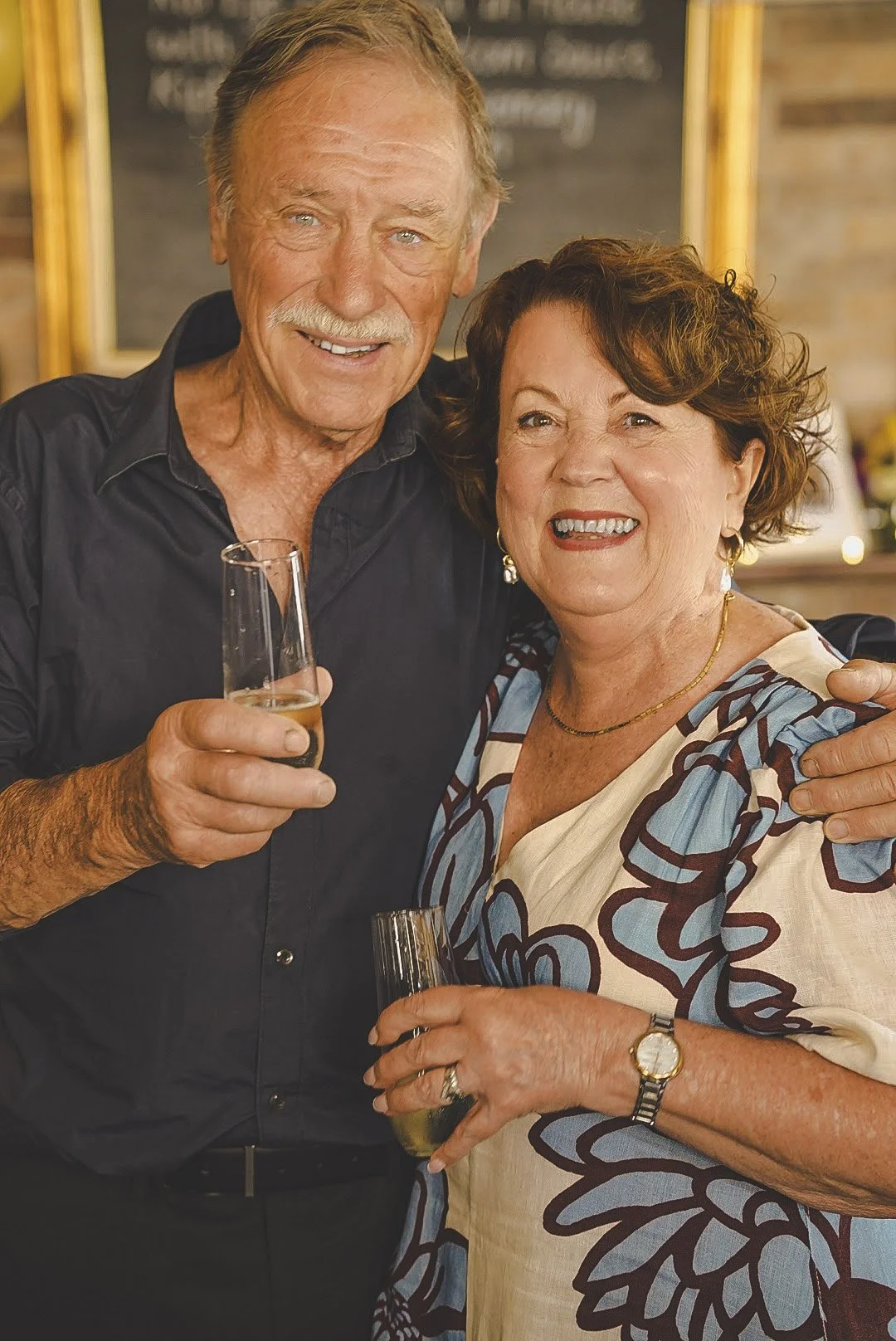 An older man and woman smiling and holding drinks at a social gathering, with a chalkboard menu in the background.