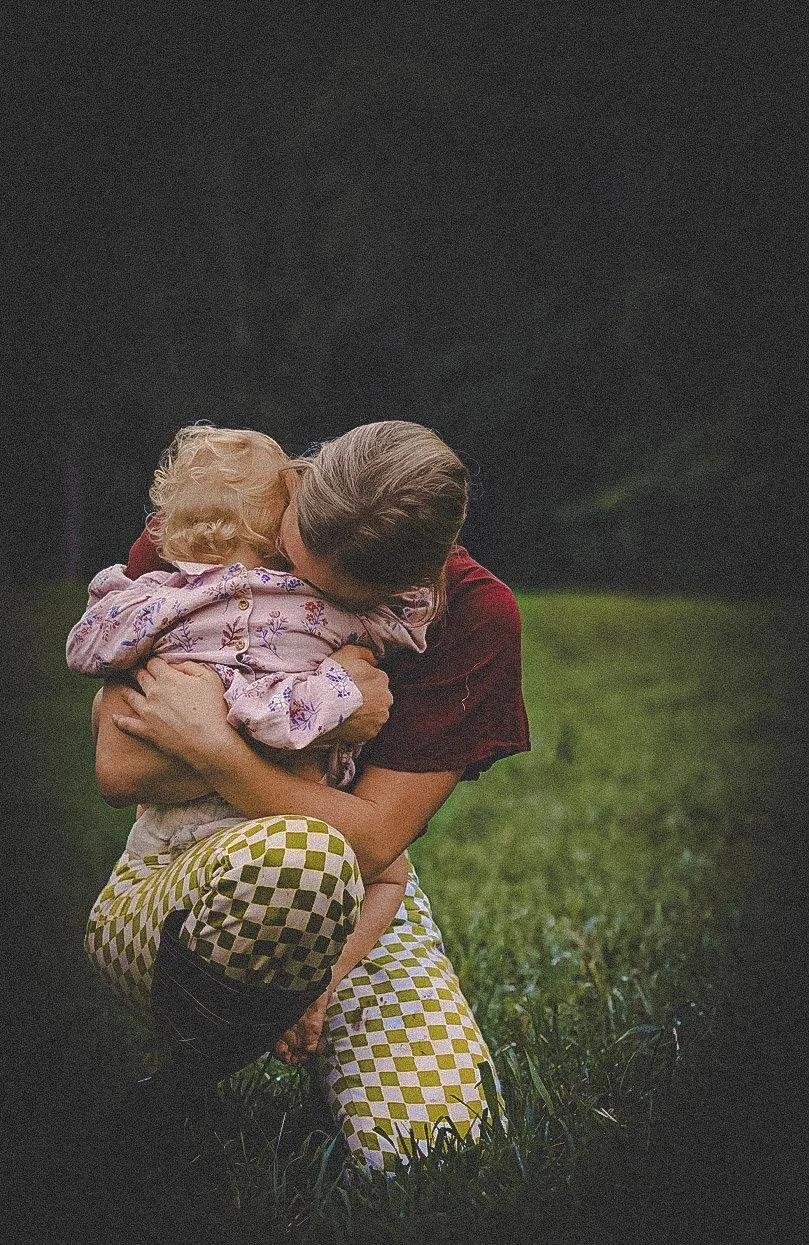 A woman crouching in a field hugging a young child, both with their faces pressed together, in an outdoor setting with grass and trees in the background.