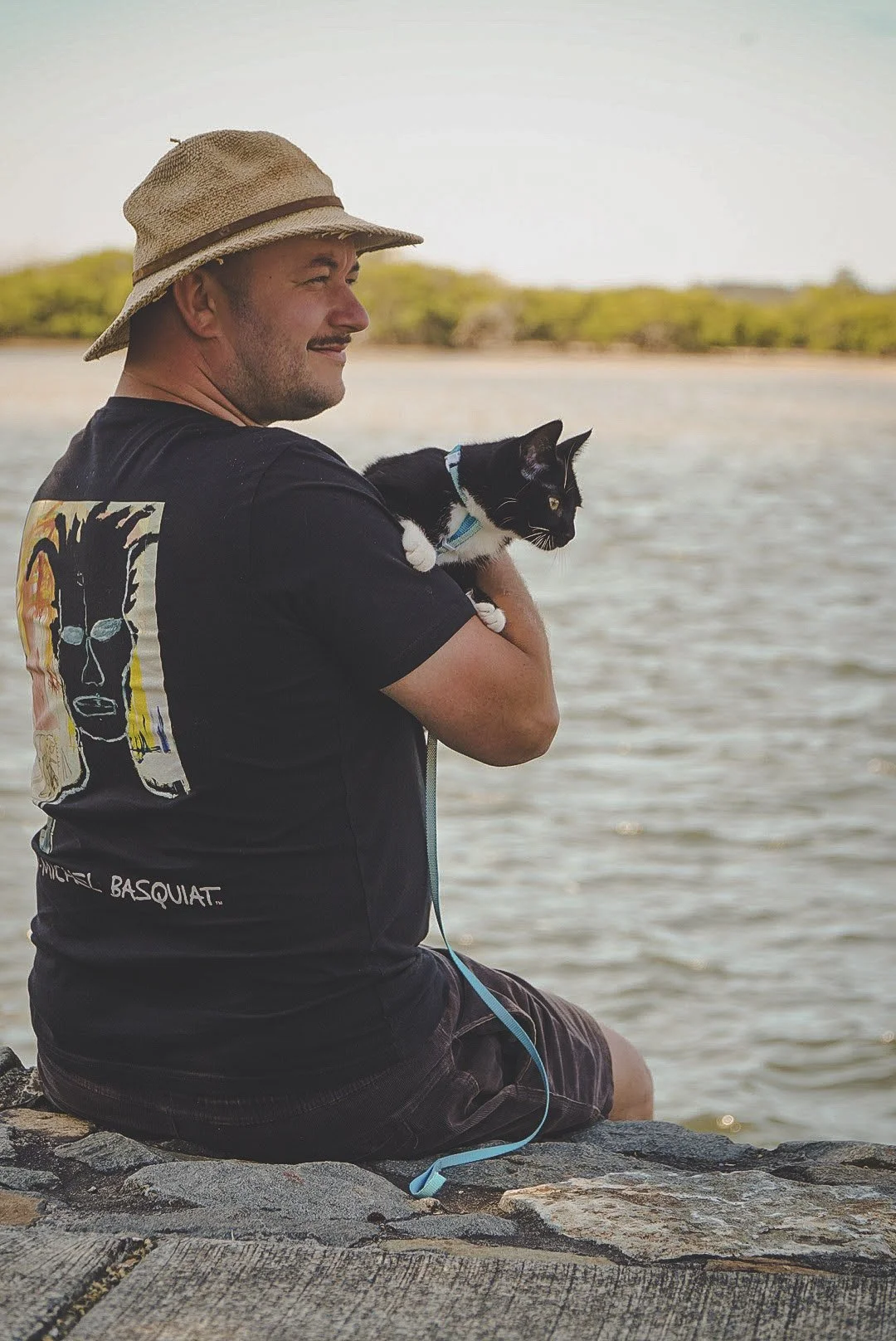 A man sitting on a rock by the water holding a black and white cat, wearing a straw hat and a black t-shirt with a colorful graphic on the back