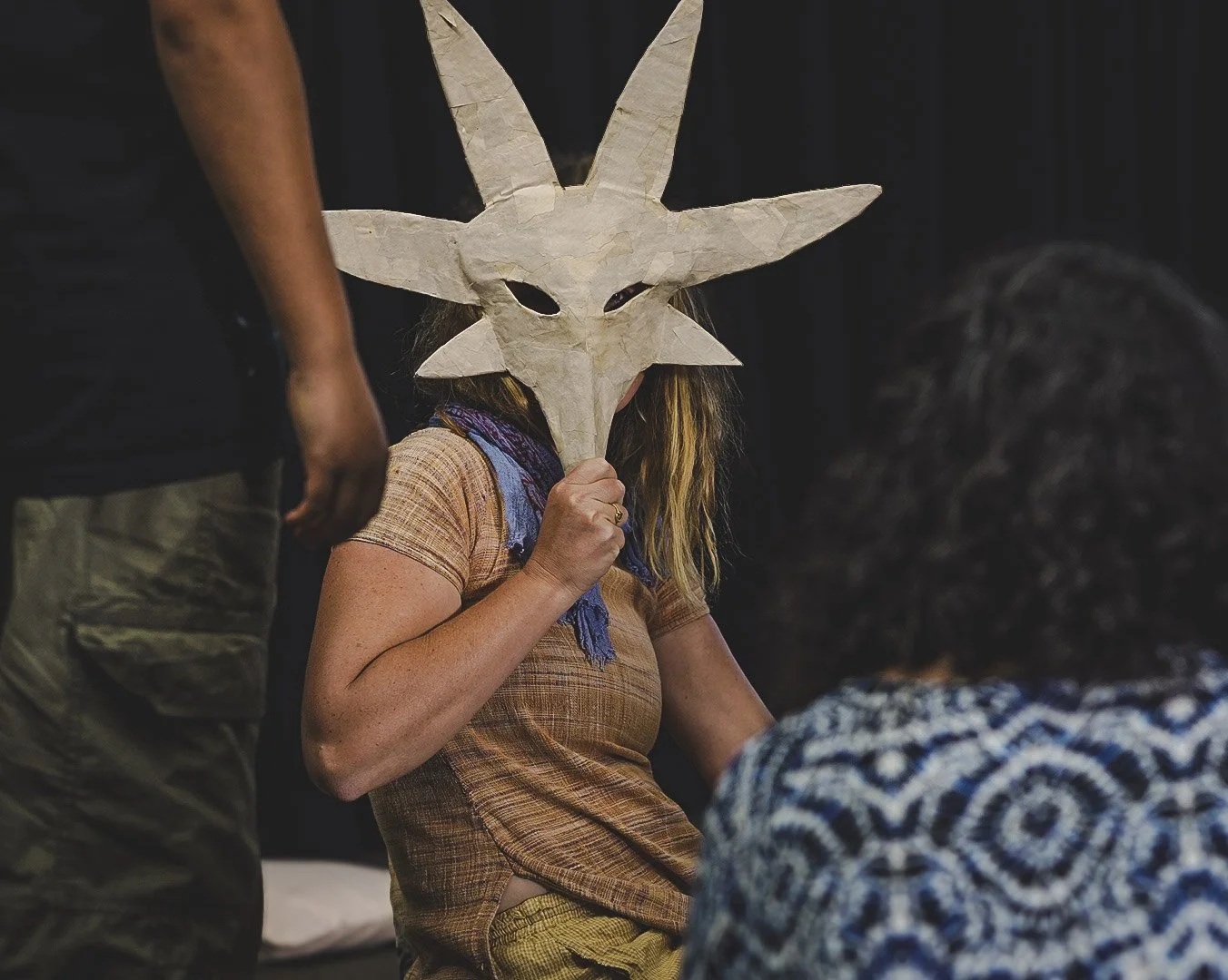 Person wearing a handmade paper mask resembling a creature with large ears and elongated face, sitting indoors near a group of people.