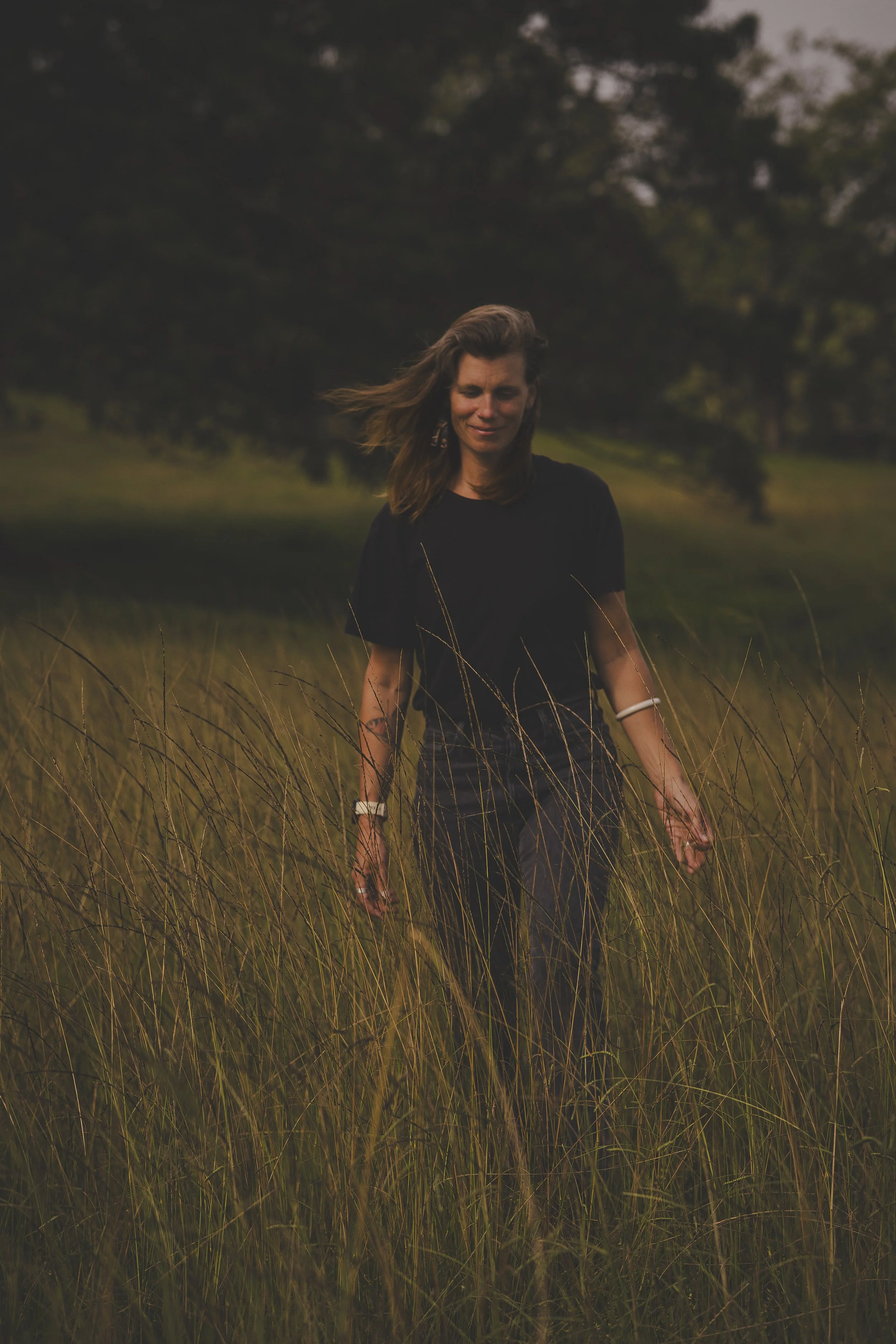 A woman with shoulder-length hair walking through a grassy field during sunset or dusk, wearing black clothing and a white bracelet, with trees in the background.