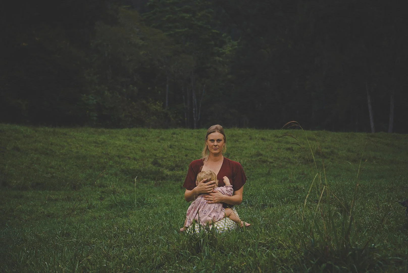 Woman sitting on grassy field holding a young child close to her chest with a forested background.
