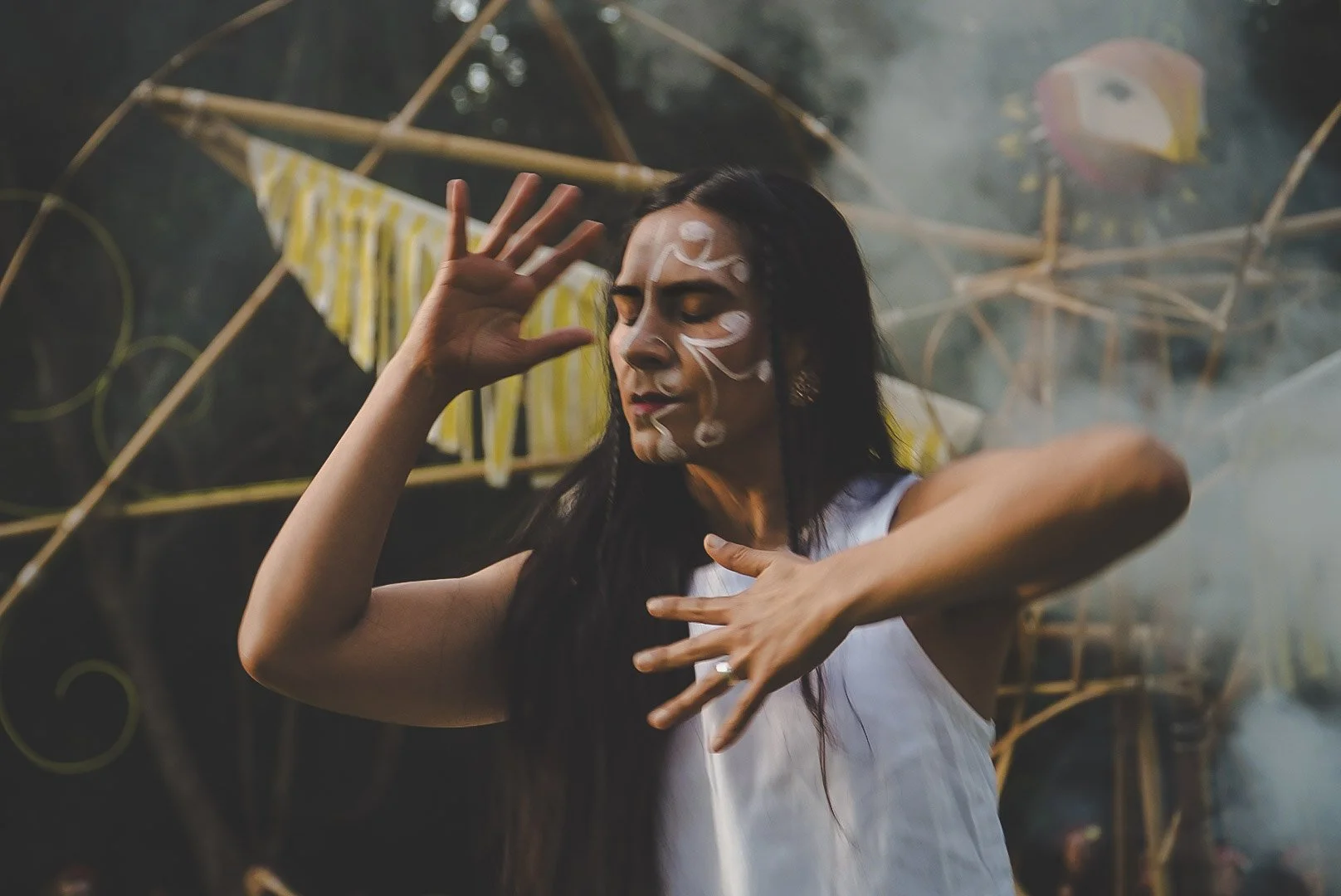 A woman dancing outdoors with white face paint designs, wearing a white dress, and surrounded by bamboo structures and colorful decorations.