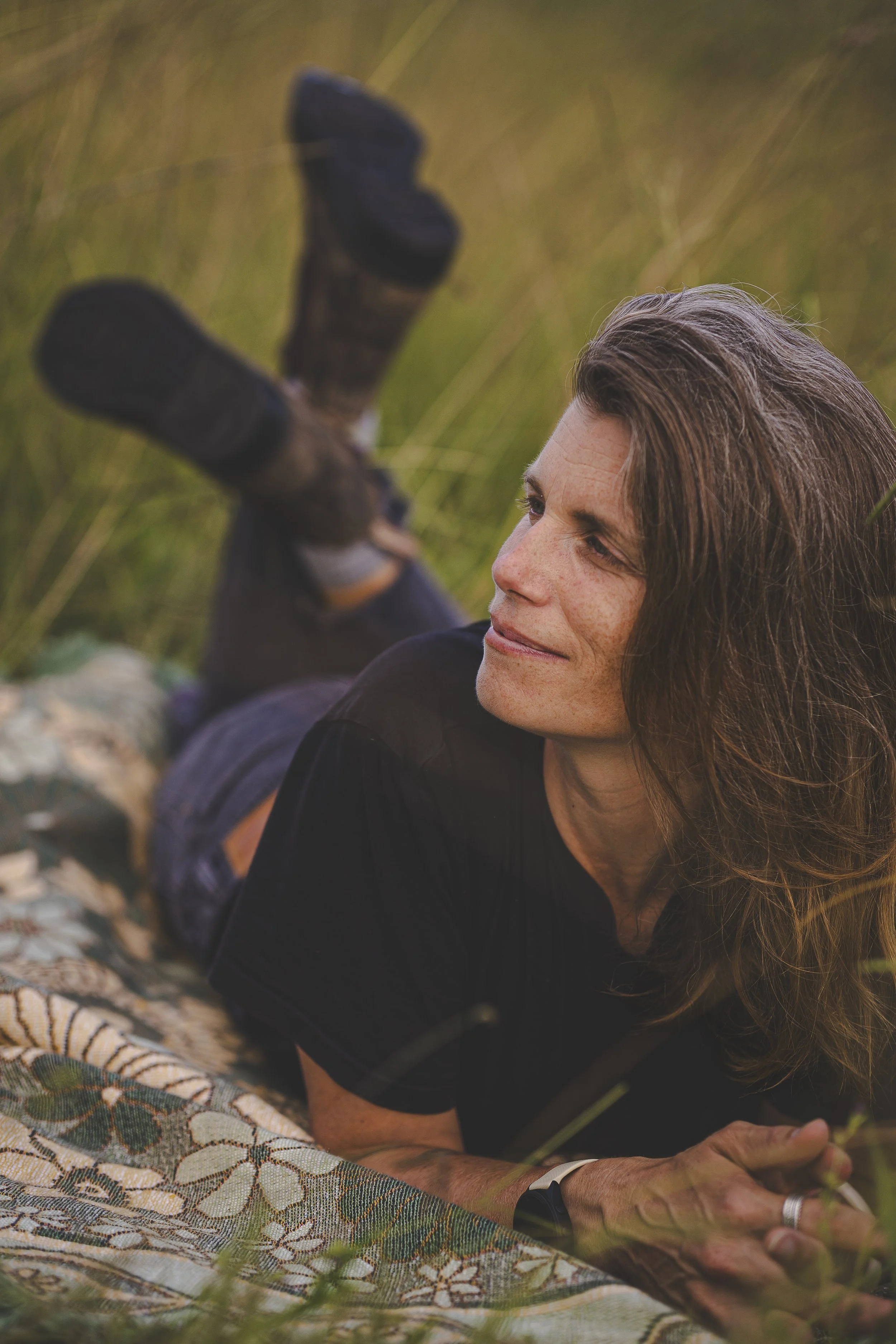 A woman with long brown hair relaxing outdoors on a patterned blanket in a grassy field, looking peacefully to the side.