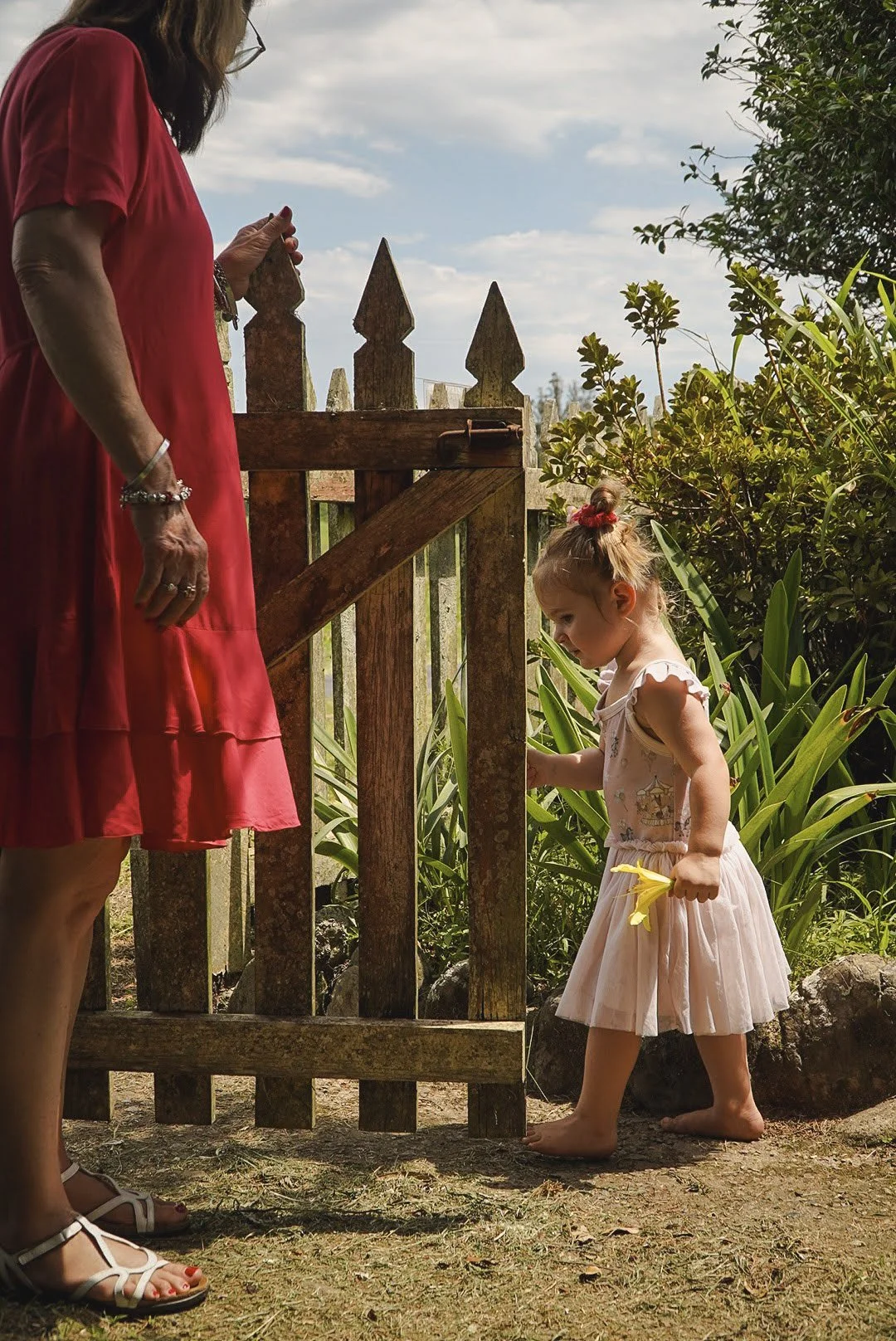 A woman in a red dress and white sandals standing beside a young girl in a pink dress, holding a yellow flower, as they stand near a wooden gate in a garden with greenery and plants.