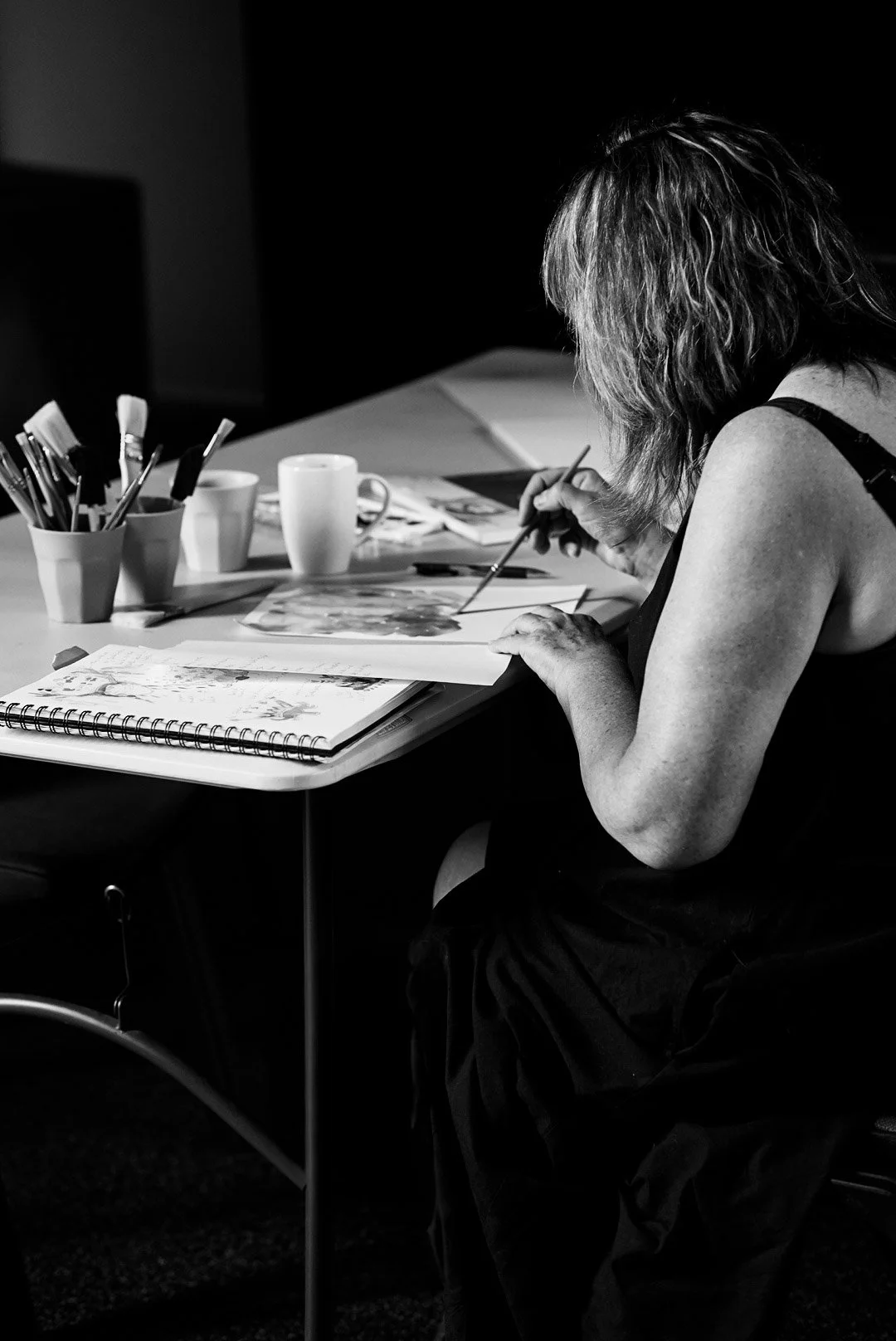 A woman sitting at a table, painting or drawing on a piece of paper, surrounded by art supplies and mugs in a black-and-white photo.