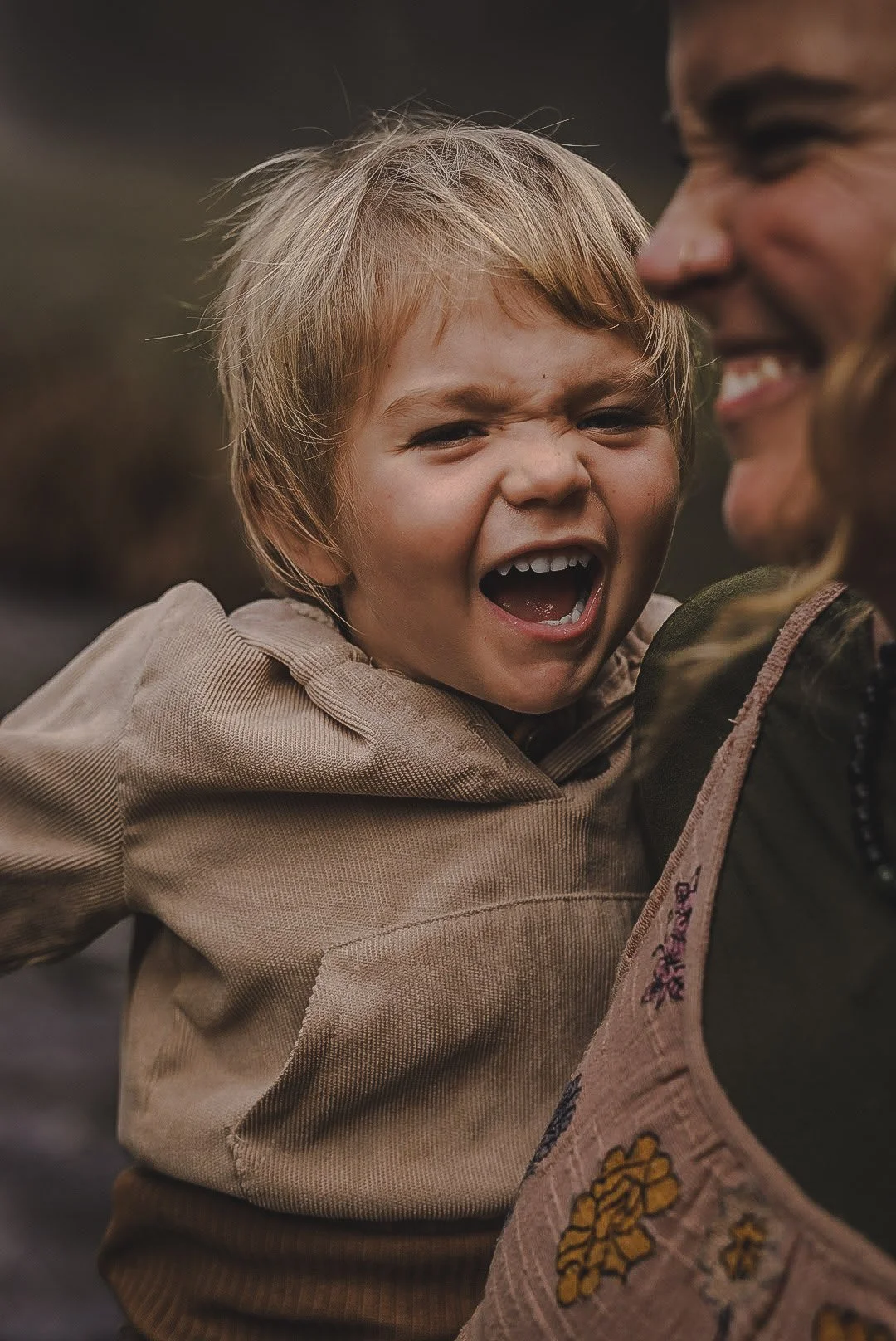 A young boy with blond hair and a tan jacket laughing with a woman, both showing joyful expressions.