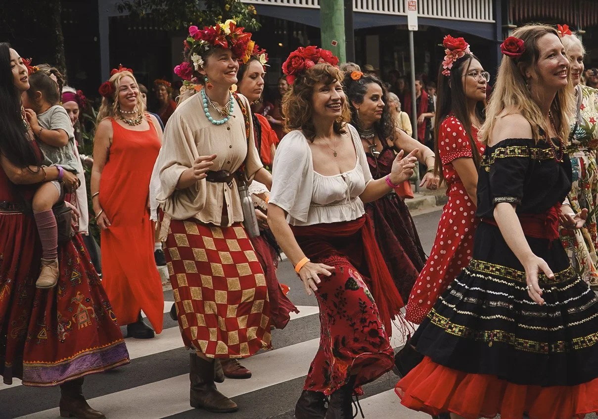 Women dancing in a parade, wearing colorful traditional Mexican dresses and floral headpieces.