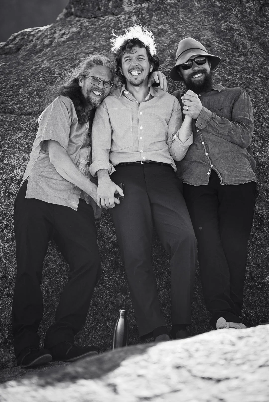 Three men smiling and holding hands, standing against a rocky background, with one wearing glasses, another with curly hair, and the third with a wide-brimmed hat and sunglasses.