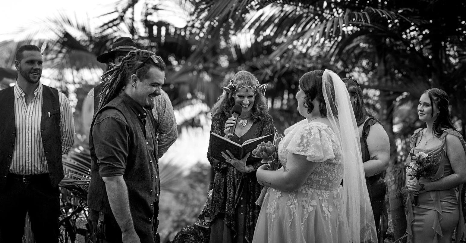 A wedding ceremony outdoors with the bride and groom facing each other while the officiant reads. The officiant is a woman with curly hair and wears a floral crown, holding a microphone and a book. The bride is in a white dress with lace, holding a b