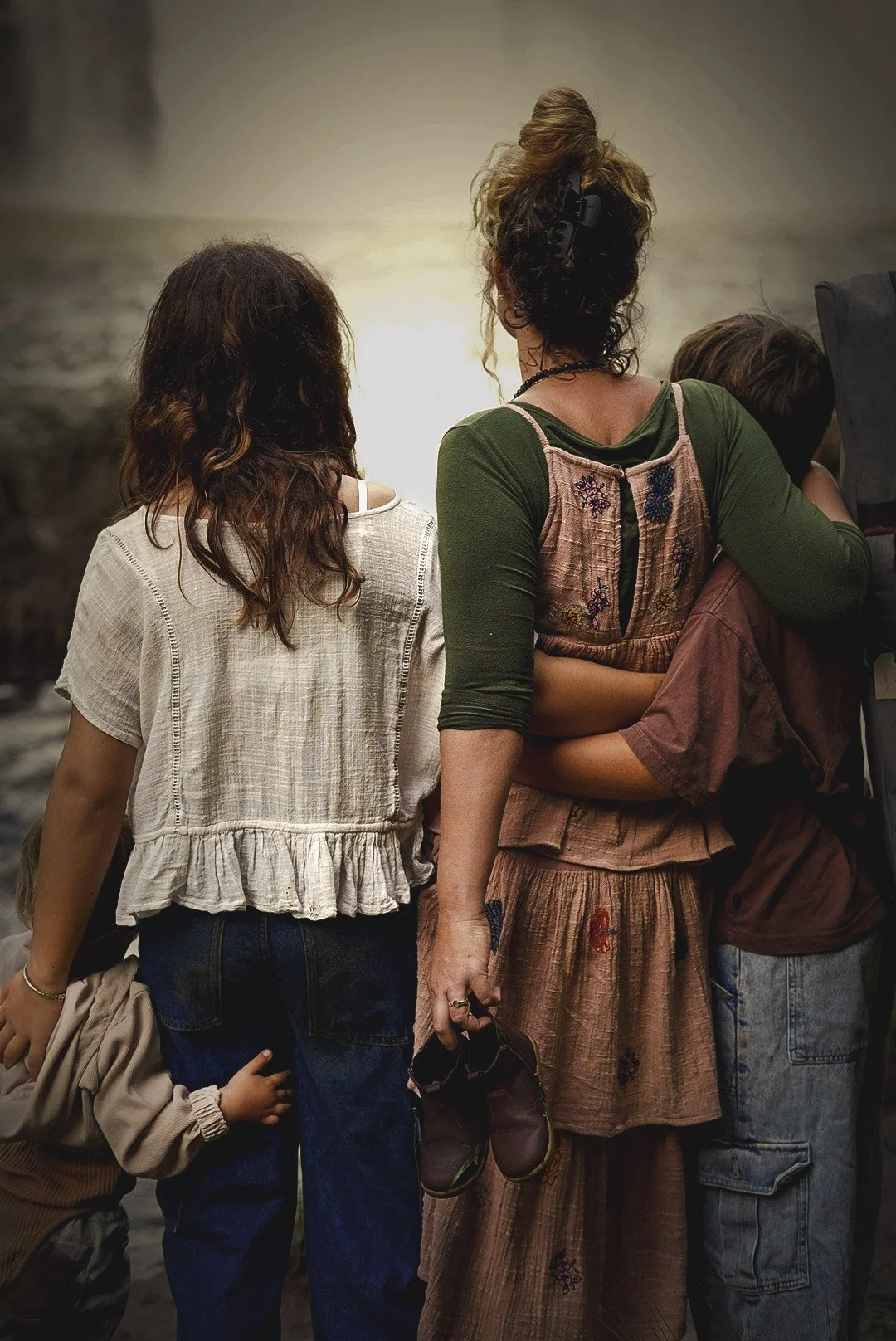 People standing outdoors during sunset, viewed from the back, with one woman holding a child's hand and another woman holding shoes.