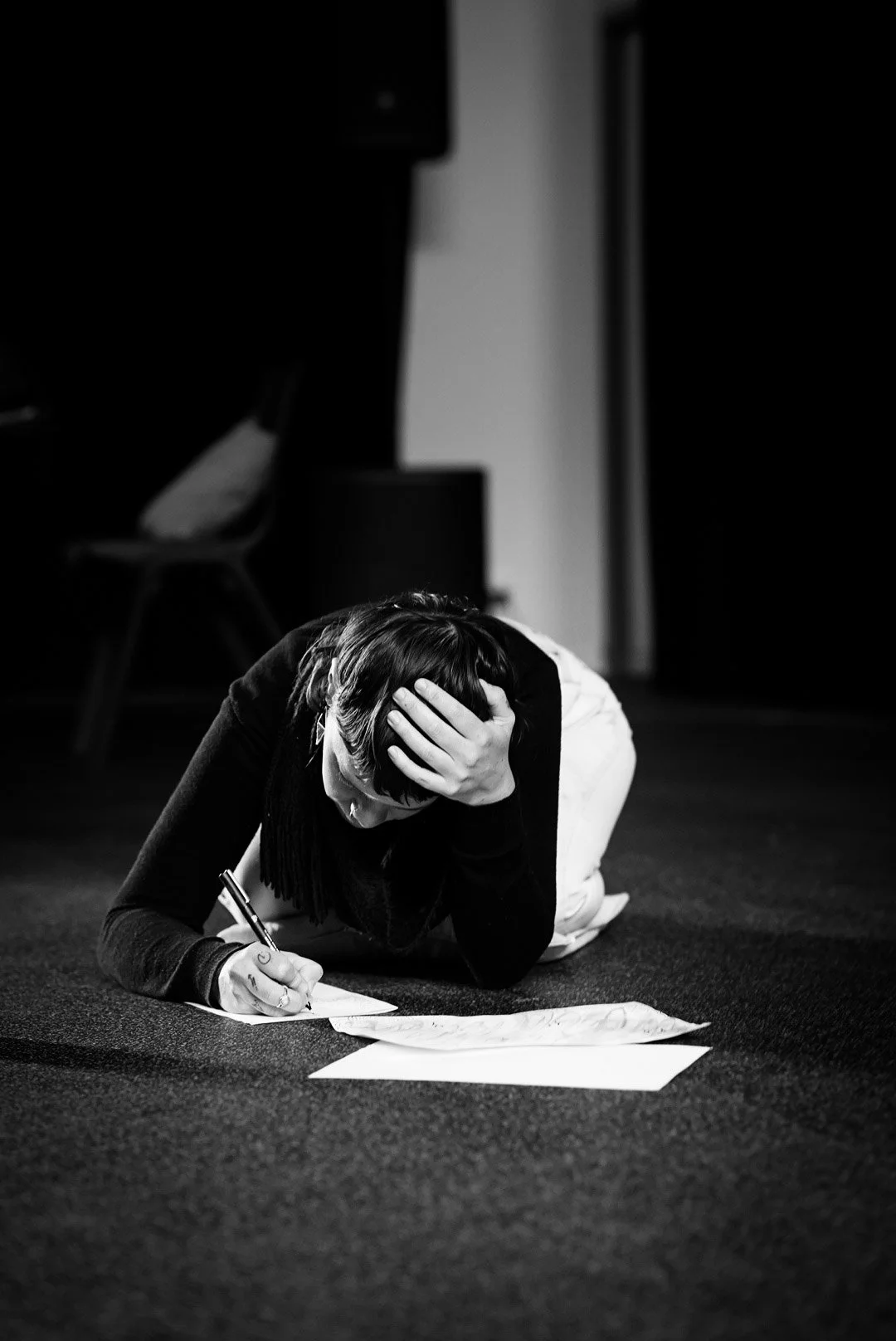 A woman lying on the floor, clutching her head with one hand and writing on paper with the other, appears distressed in a black-and-white photo.