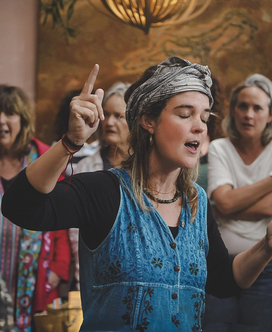 A woman with a headscarf and blue embroidered dress is raising her hand with her eyes closed, appearing to be singing or praying. Several women are in the background, some with arms crossed or looking attentively.
