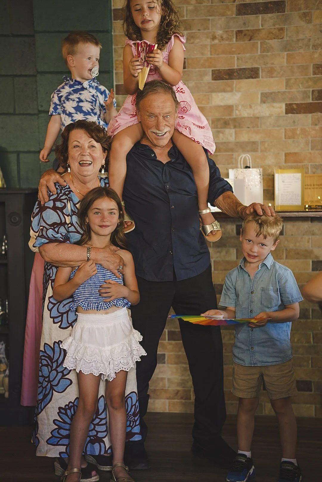 A family celebrating a special occasion with many generations. Six people are posed in front of a brick wall. The elderly man is in the middle, smiling, with a woman and children around him. The woman is wearing a colorful dress, and the young girl i