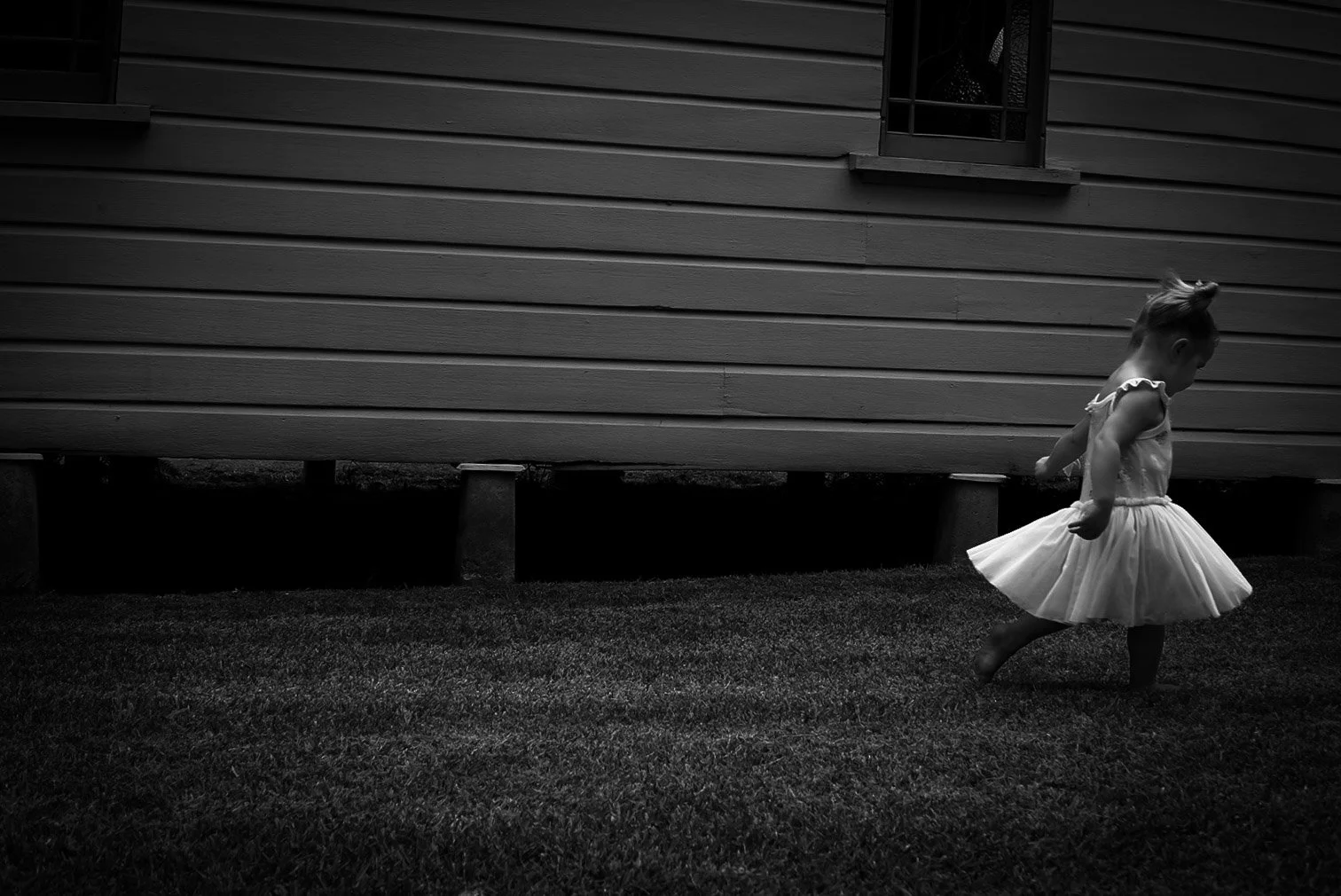 A young girl in a light-colored dress bouncing or spinning on the grass in front of a wooden house, with a small window visible in the background.