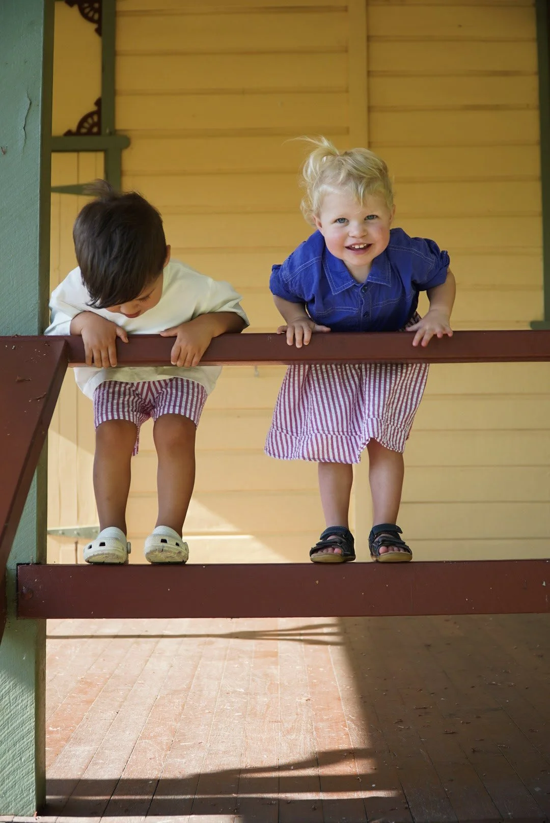 Two young children peering over the railing on a wooden porch, smiling and playing.
