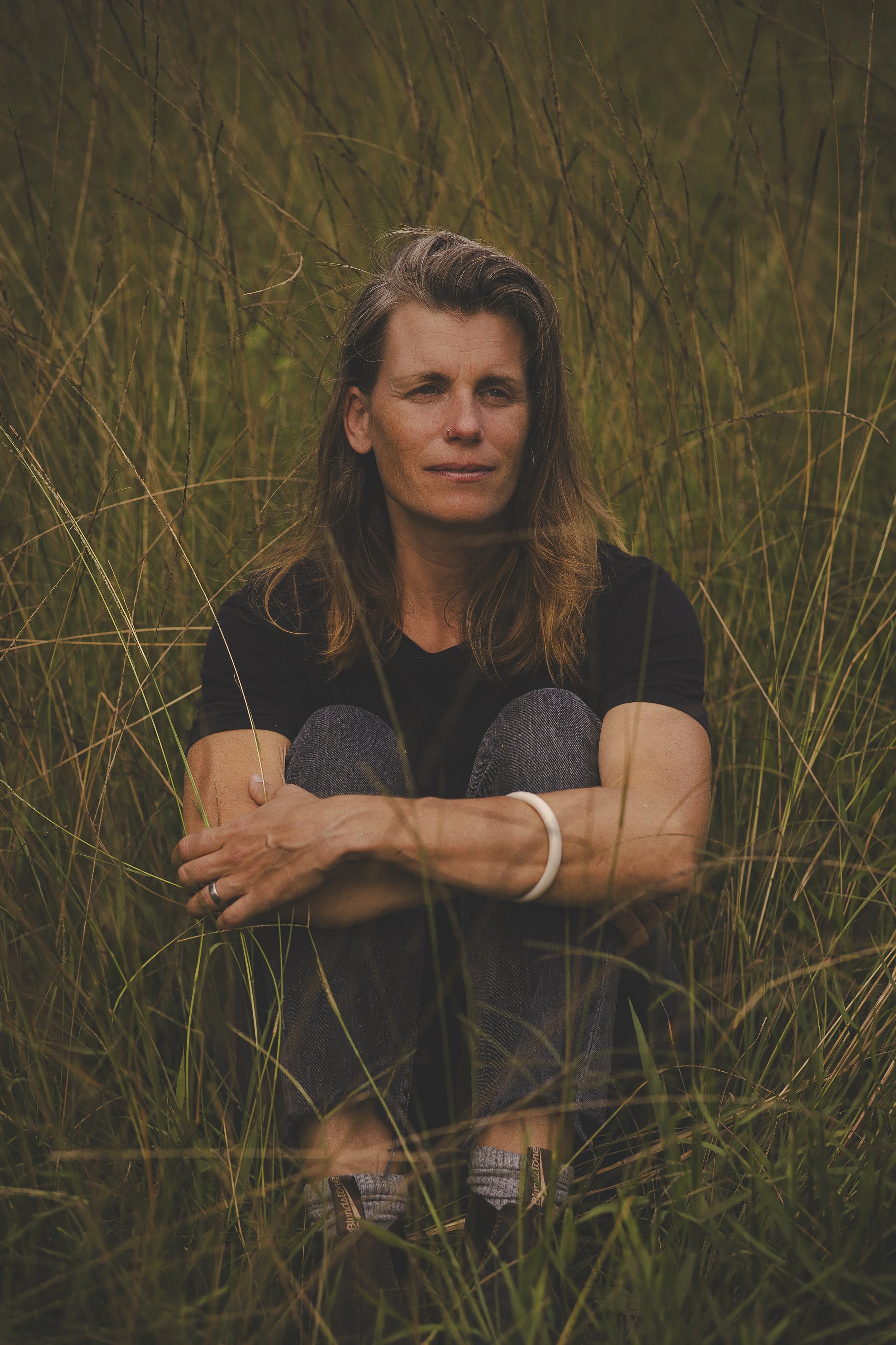 A woman sitting among tall grass, with a contemplative expression, wearing a black shirt, jeans, and sneakers, in an outdoor grassy field.