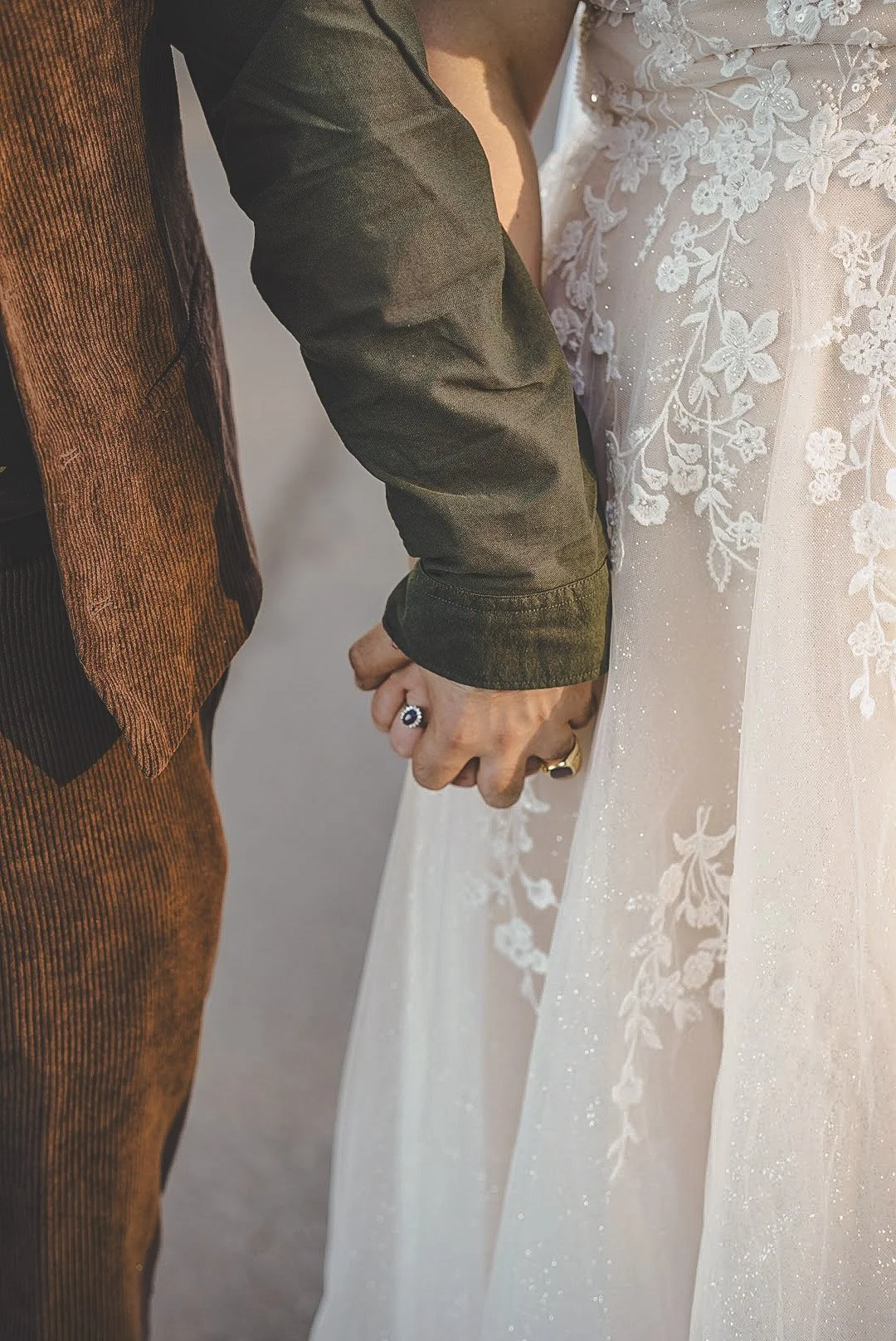 Couple holding hands during a wedding ceremony, with the bride wearing a white lace dress and the groom in a dark jacket and brown pants.