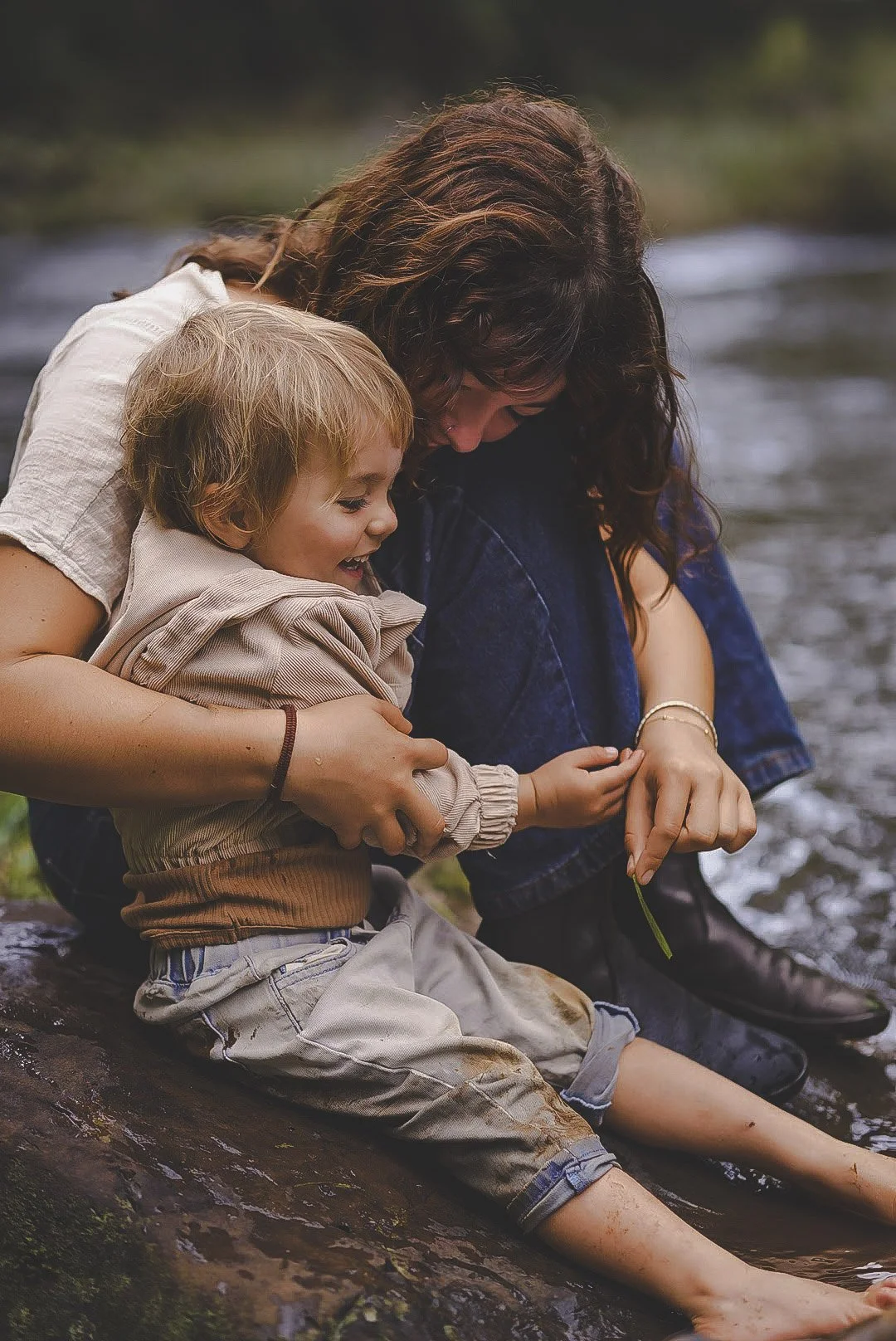 Mother and a young boy are sitting on a rock near a river, enjoying a moment together, with the woman helping the boy touch the water.