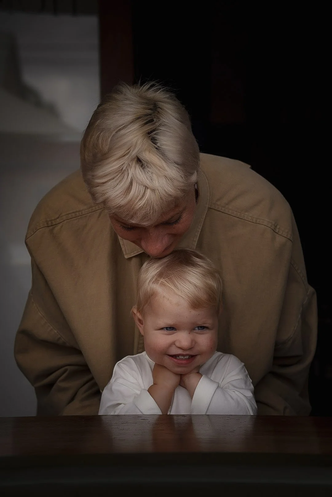 A man with blonde hair leaning down towards a young blonde-haired child who is smiling and resting their chin on their hands on a table.