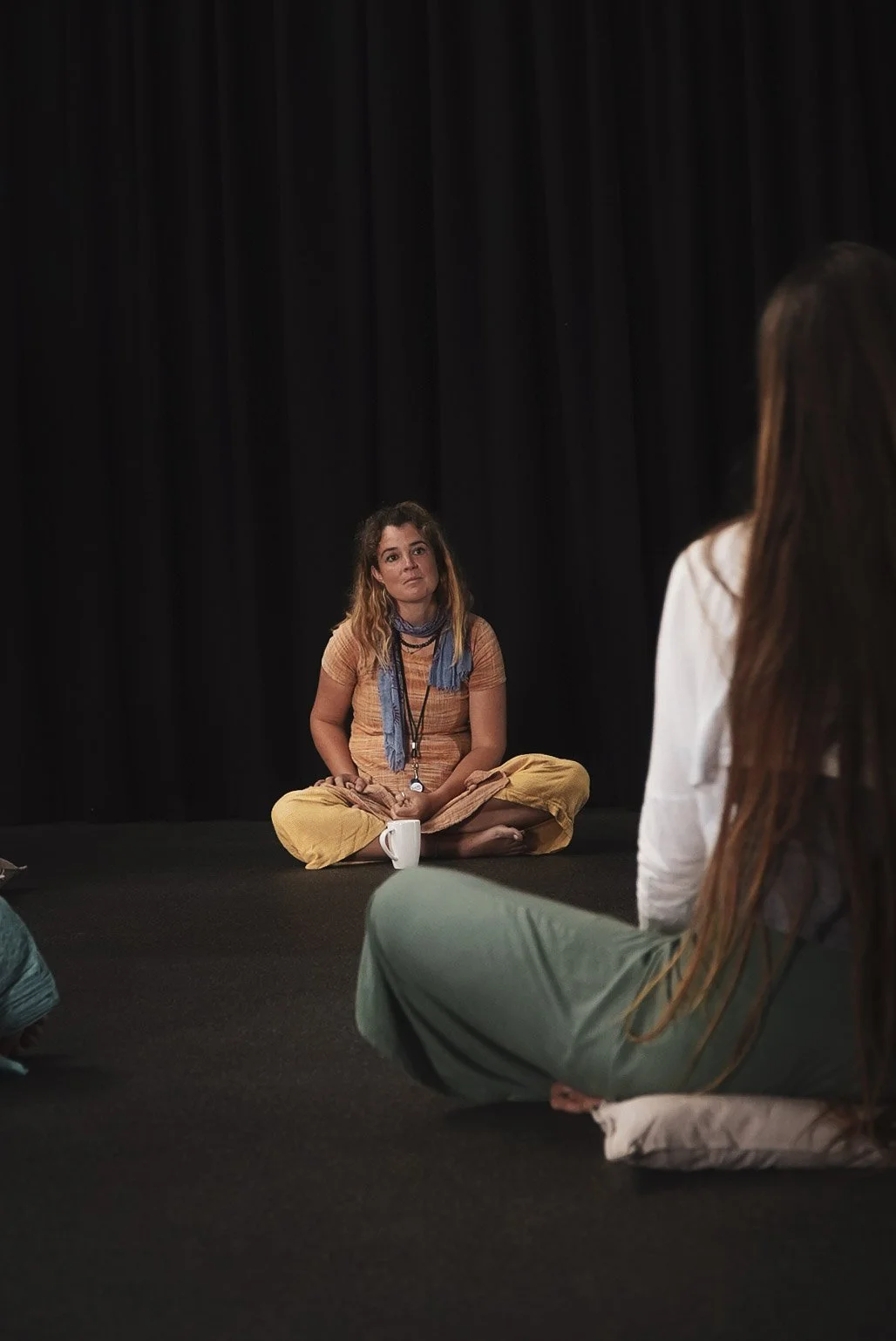 A woman with curly hair sits cross-legged on a black stage, wearing a mustard-colored outfit and a blue scarf, with a white mug in front of her, as she appears to be in a discussion or listening to someone.