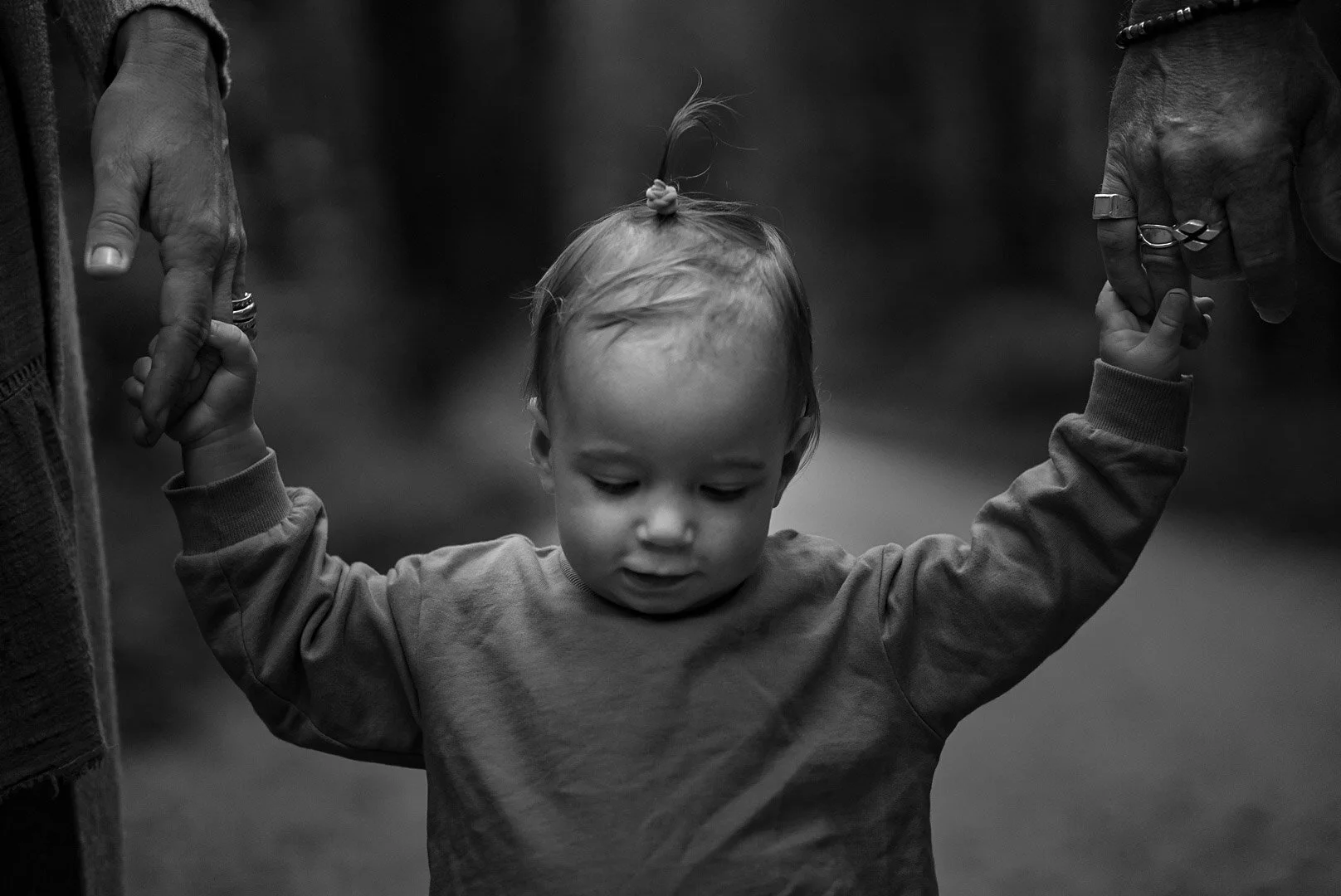 A young child with a small top knot hairstyle, holding hands with two adults, walking outdoors. The child's head is bowed and the scene is in black and white.