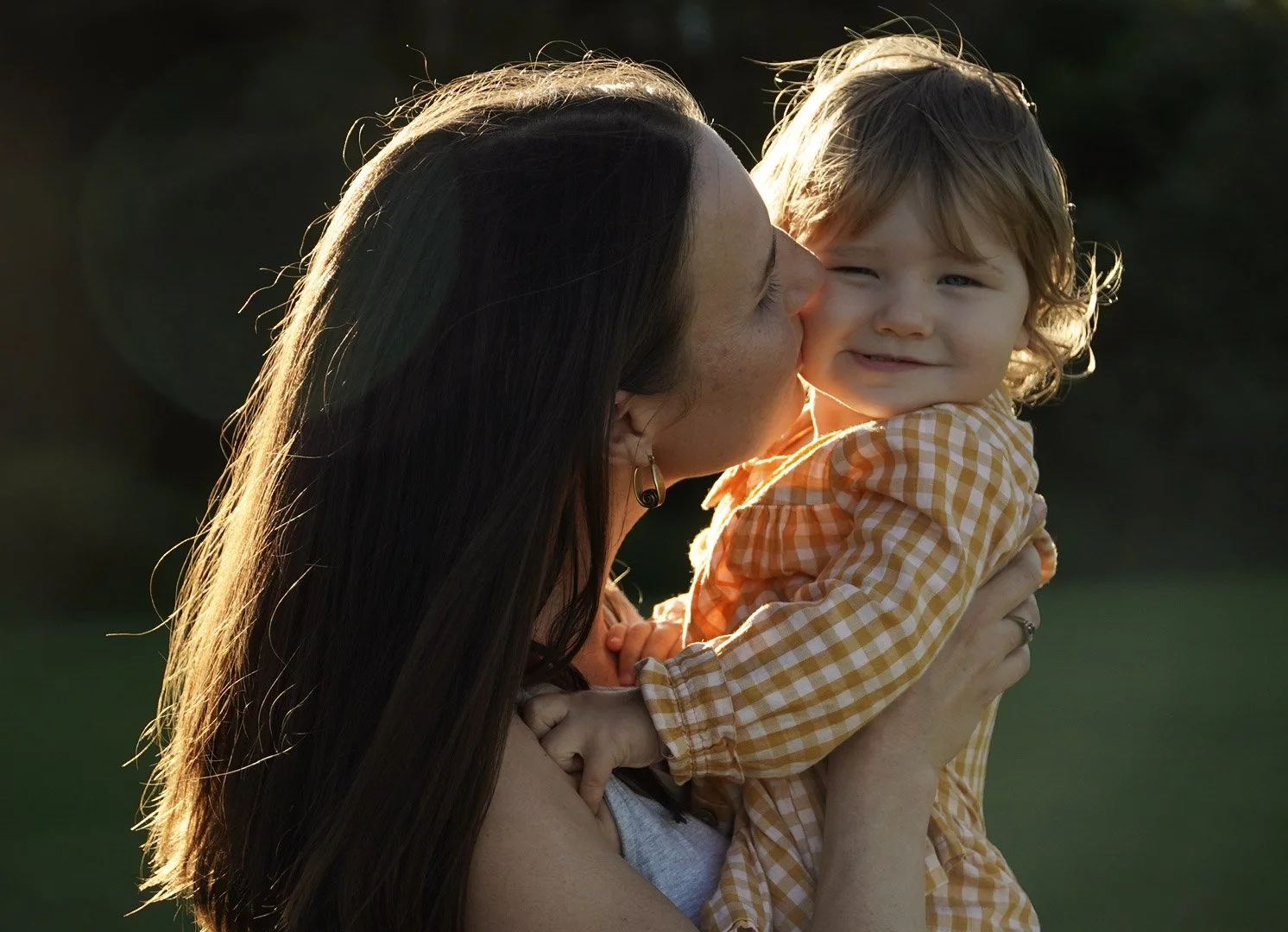 A woman kissing a smiling young child outside during sunset, with the child's face turned slightly towards the camera.
