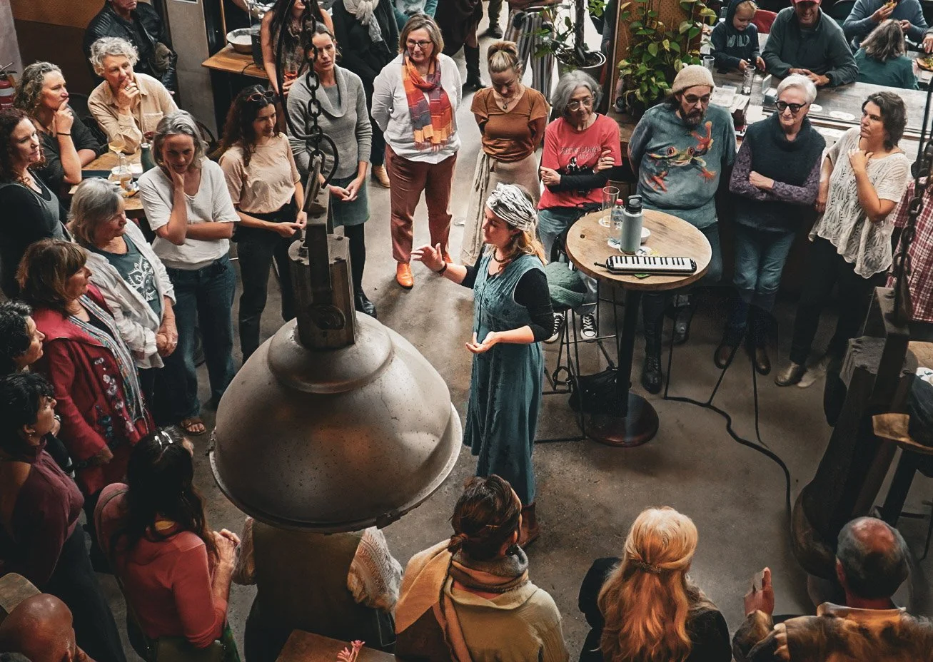 A woman with a headscarf and long shirt speaks to a crowd gathered in a cafe or restaurant. The group is listening attentively, with some standing and some seated at tables, and there is a variety of ages represented.