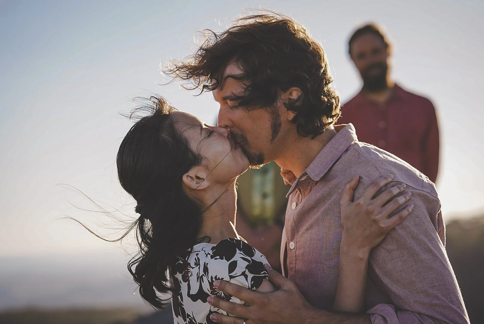 A couple kissing outdoors with a man standing in the background during sunset or sunrise.