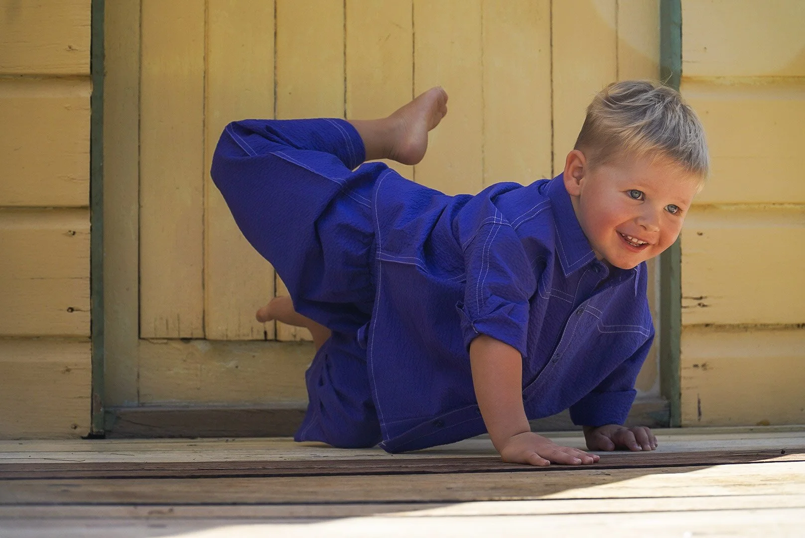 Young boy in a blue outfit crawling on a wooden porch next to a yellow house wall, smiling.